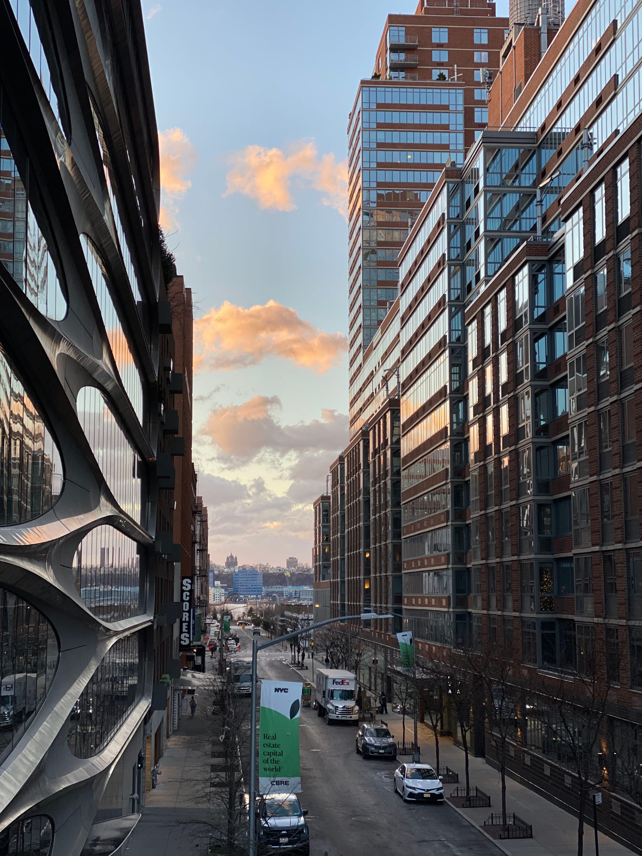 An urban street scene with tall modern buildings on both sides, a few vehicles on the road, and a sky with scattered clouds at sunset
