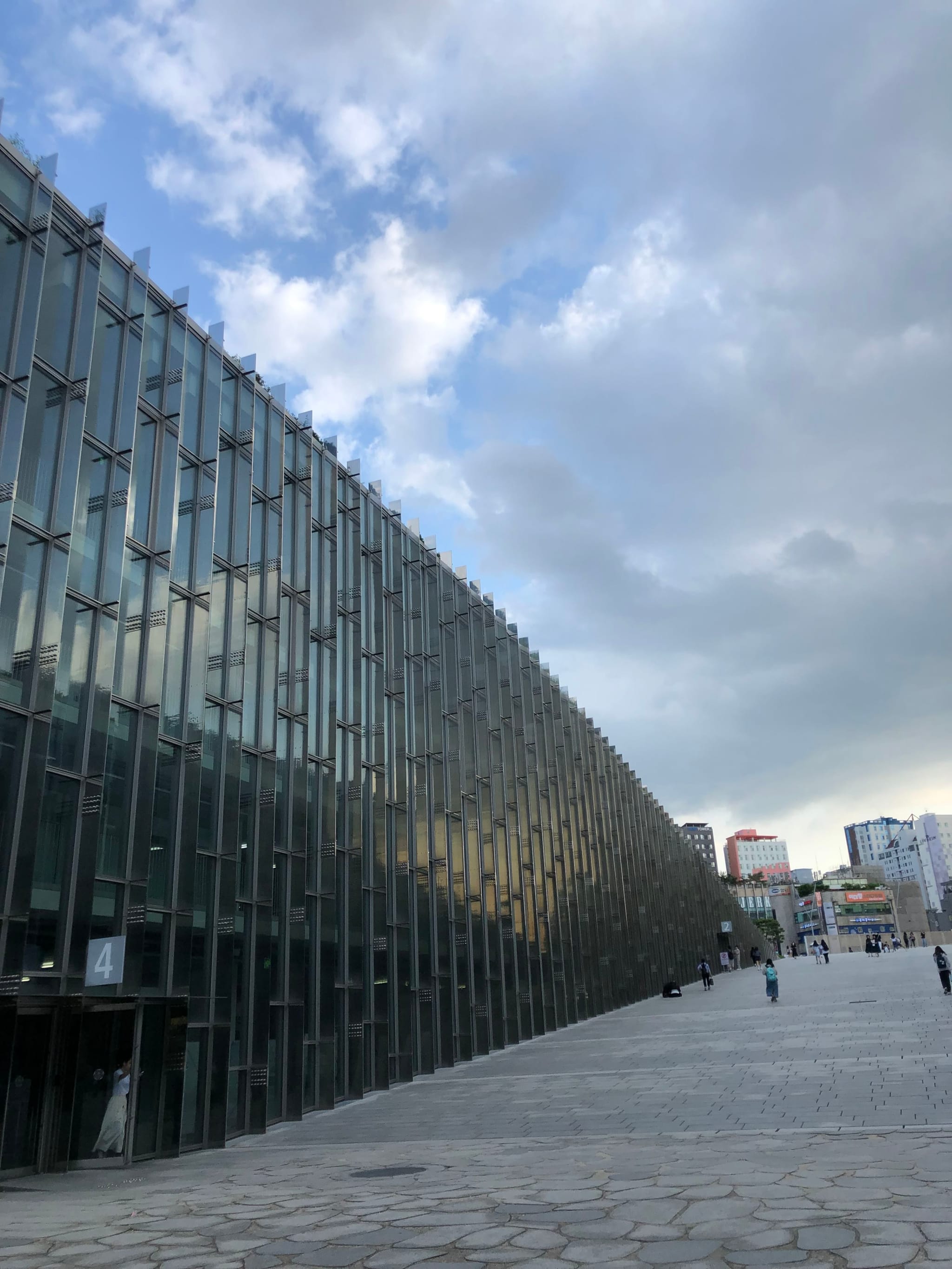 A modern glass building with a reflective facade, set against a partly cloudy sky, with a paved walkway and a few people in the distance