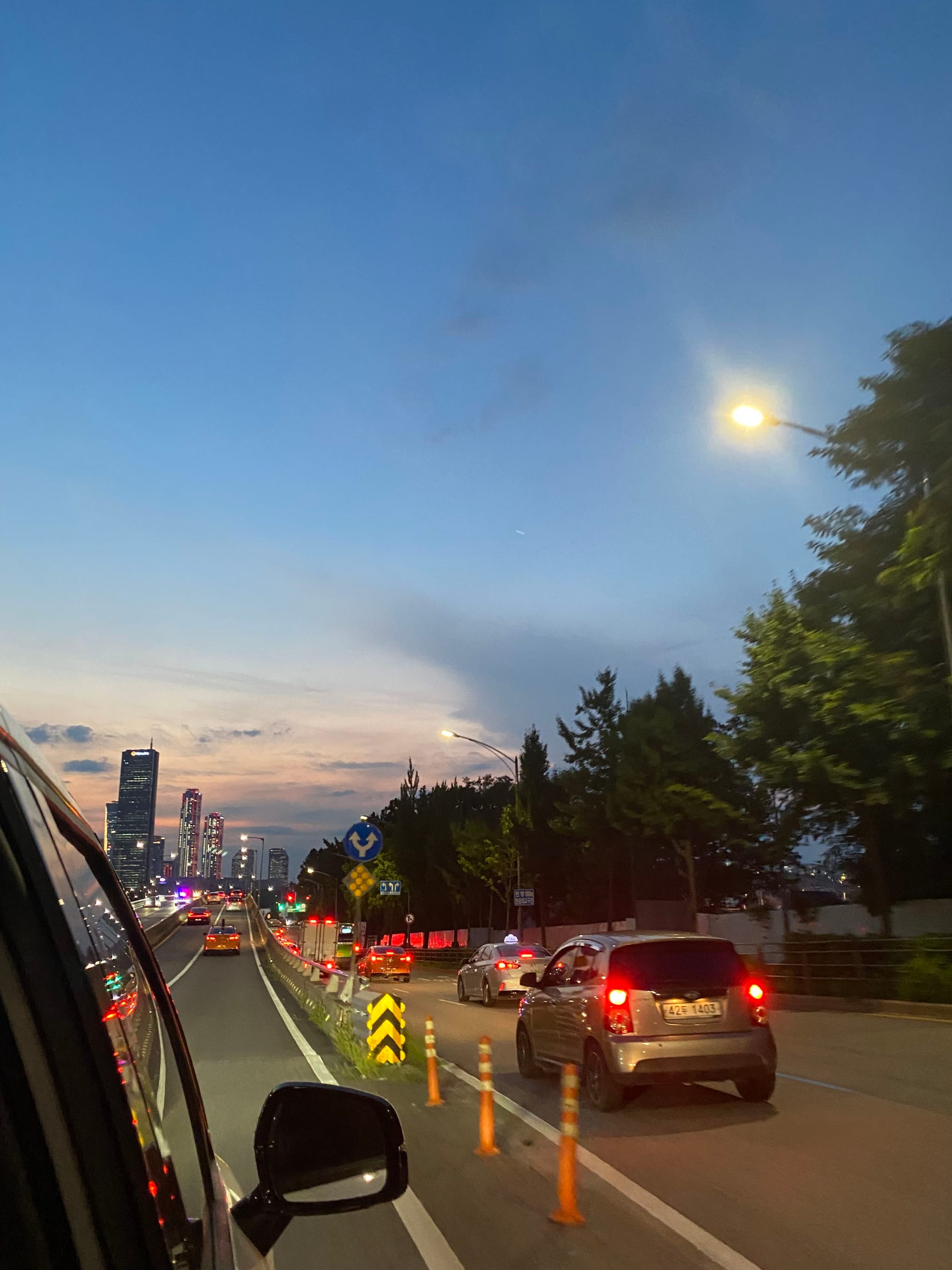 A city street at dusk with cars driving along a road lined with traffic cones. The sky is a gradient of blue and orange, and streetlights are illuminated. Trees and buildings are visible in the background