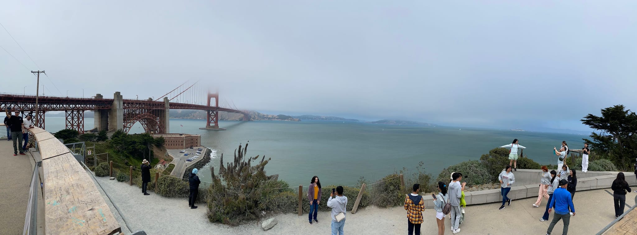 A panoramic view of the Golden Gate Bridge with people gathered at a viewpoint overlooking the water, under a cloudy sky