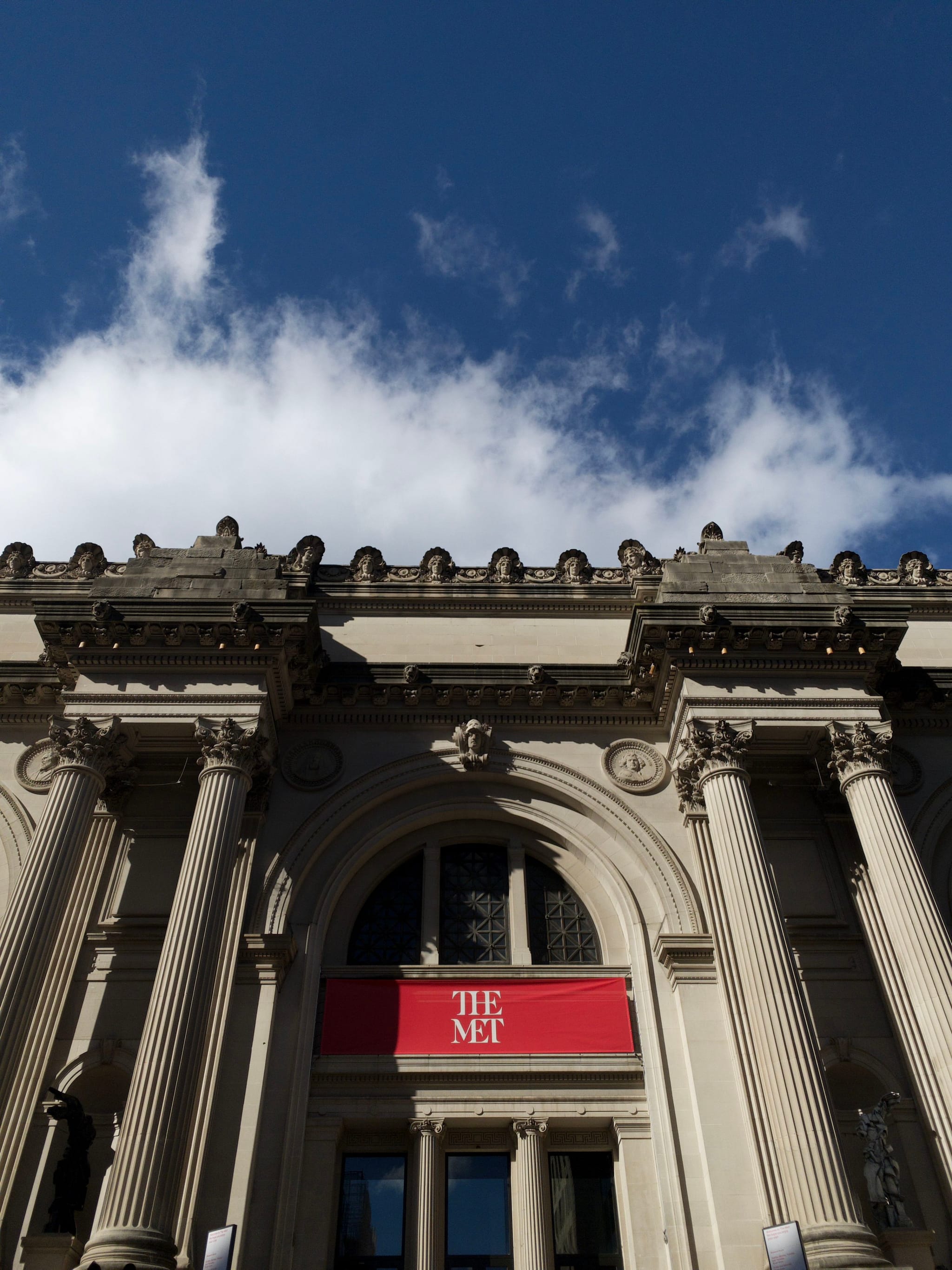 The facade of a classical building with tall columns and a red sign reading THE MET against a backdrop of a blue sky with clouds