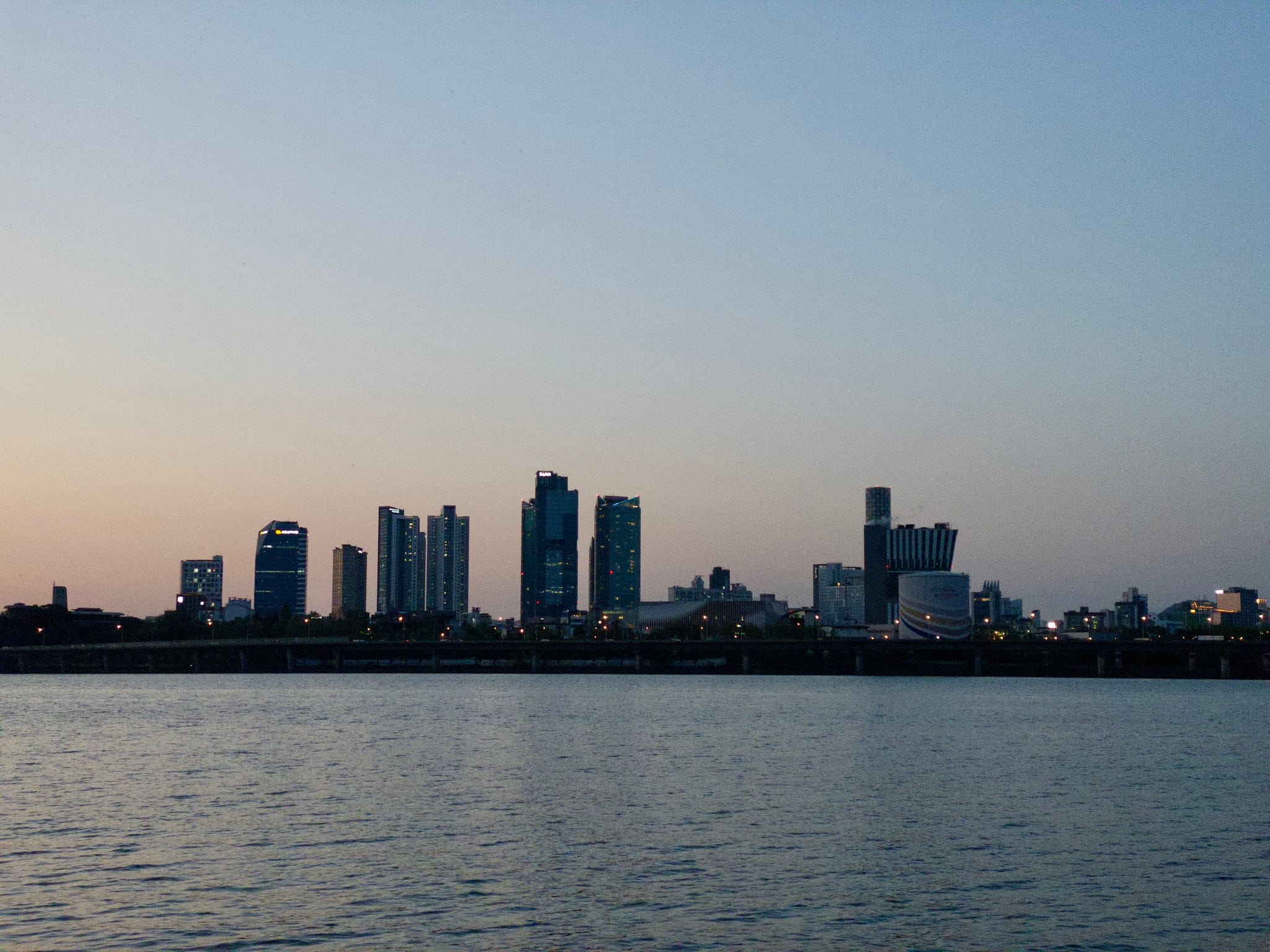 A city skyline at dusk with tall buildings silhouetted against a clear sky, reflected in a body of water in the foreground