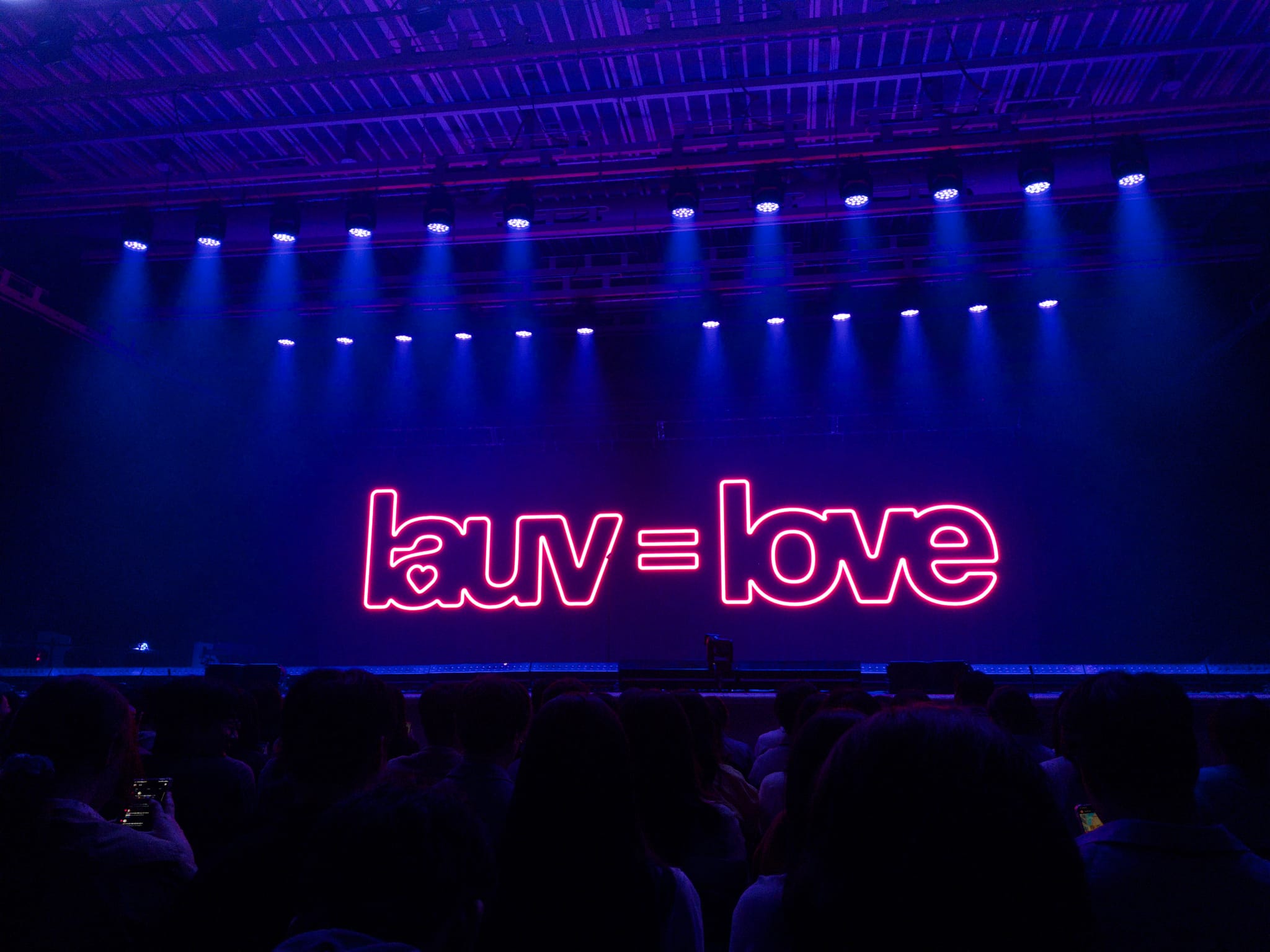 A stage with blue lighting features the phrase lauv = love in bright pink neon letters