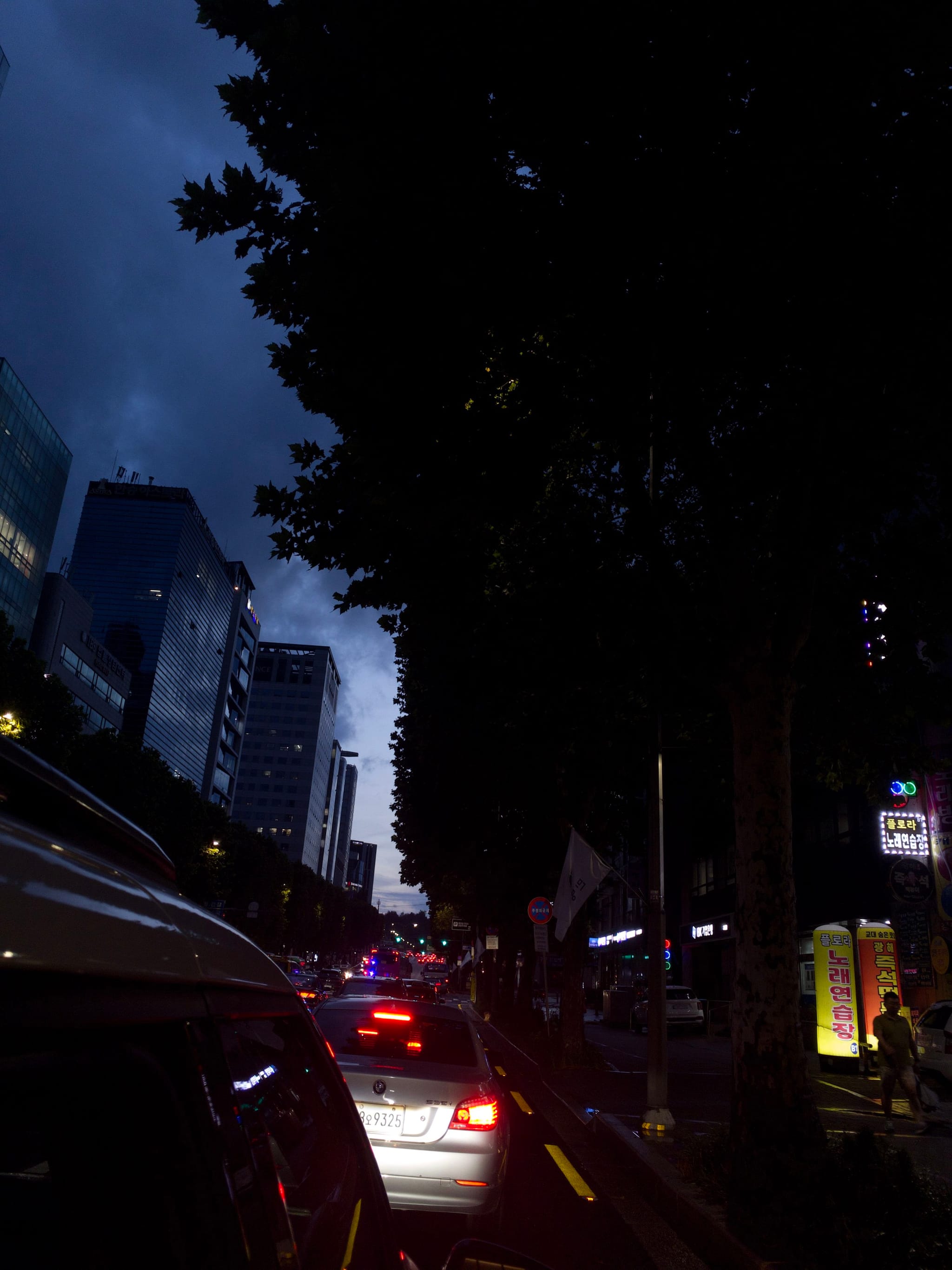 A nighttime urban street scene with cars in traffic, illuminated by headlights and taillights, flanked by tall buildings and trees