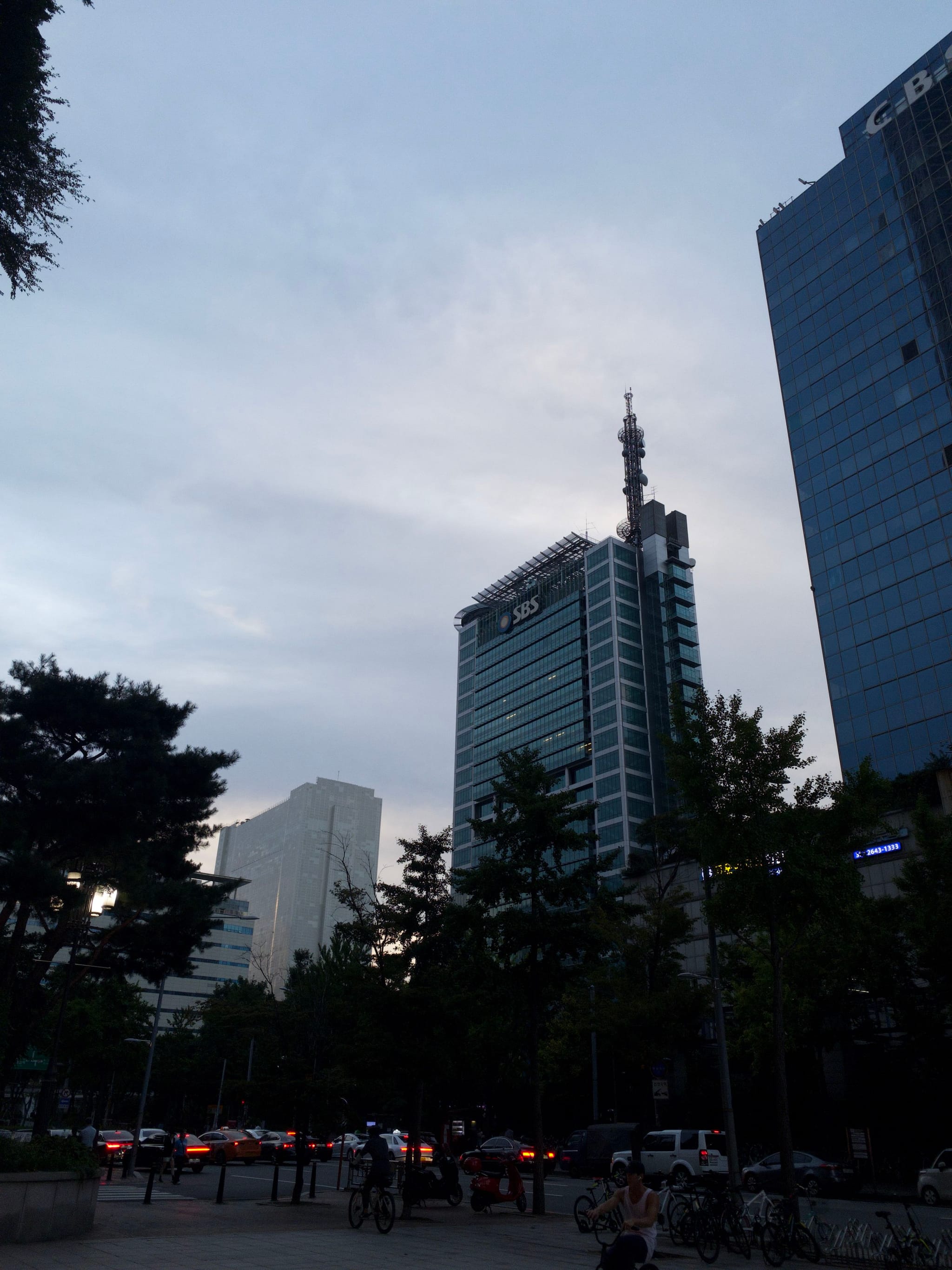 A cityscape with tall buildings, trees, and a cloudy sky, featuring a street with cars and people