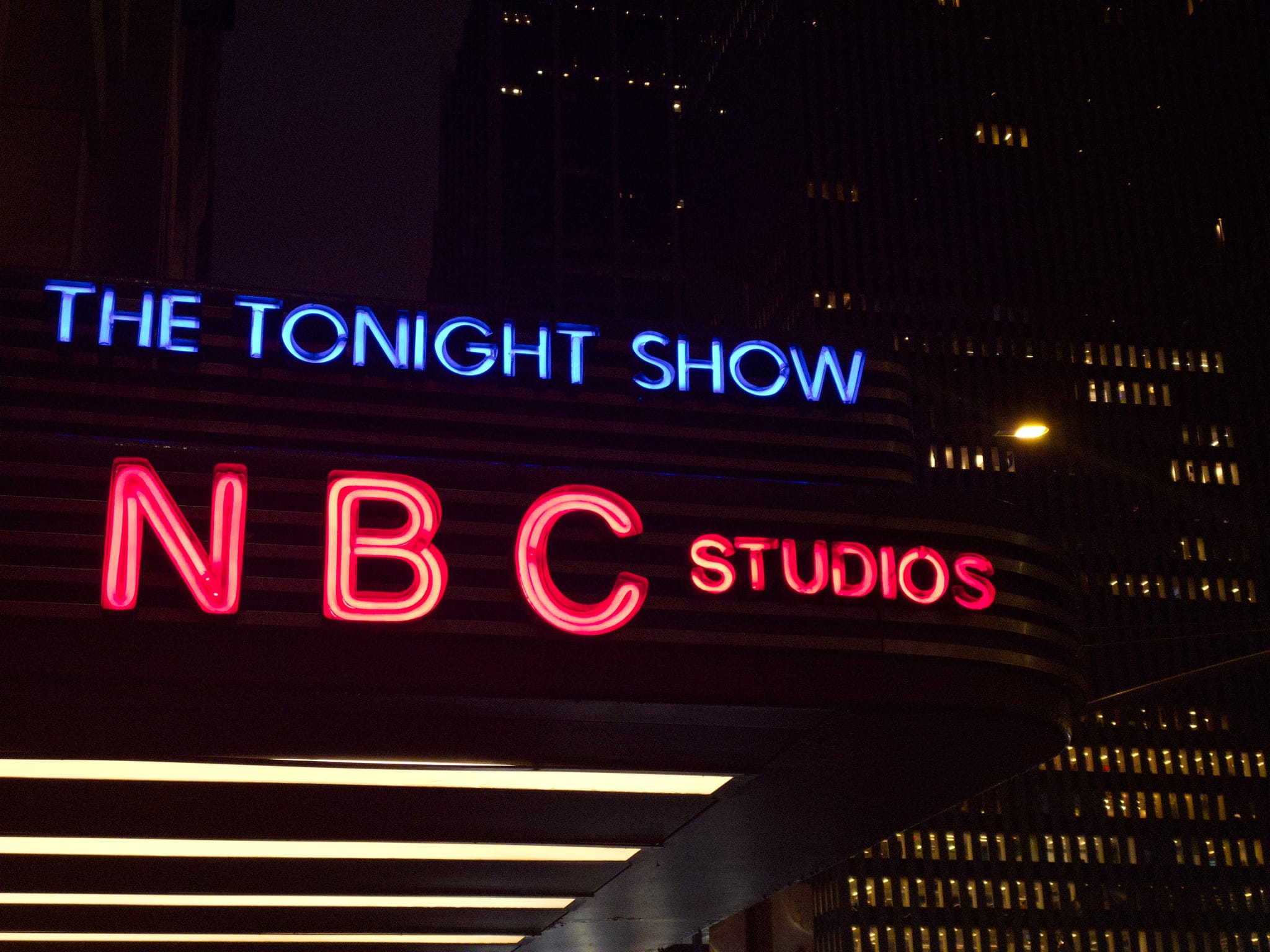A neon sign for The Tonight Show and NBC Studios with blue and red lettering, set against a dark cityscape
