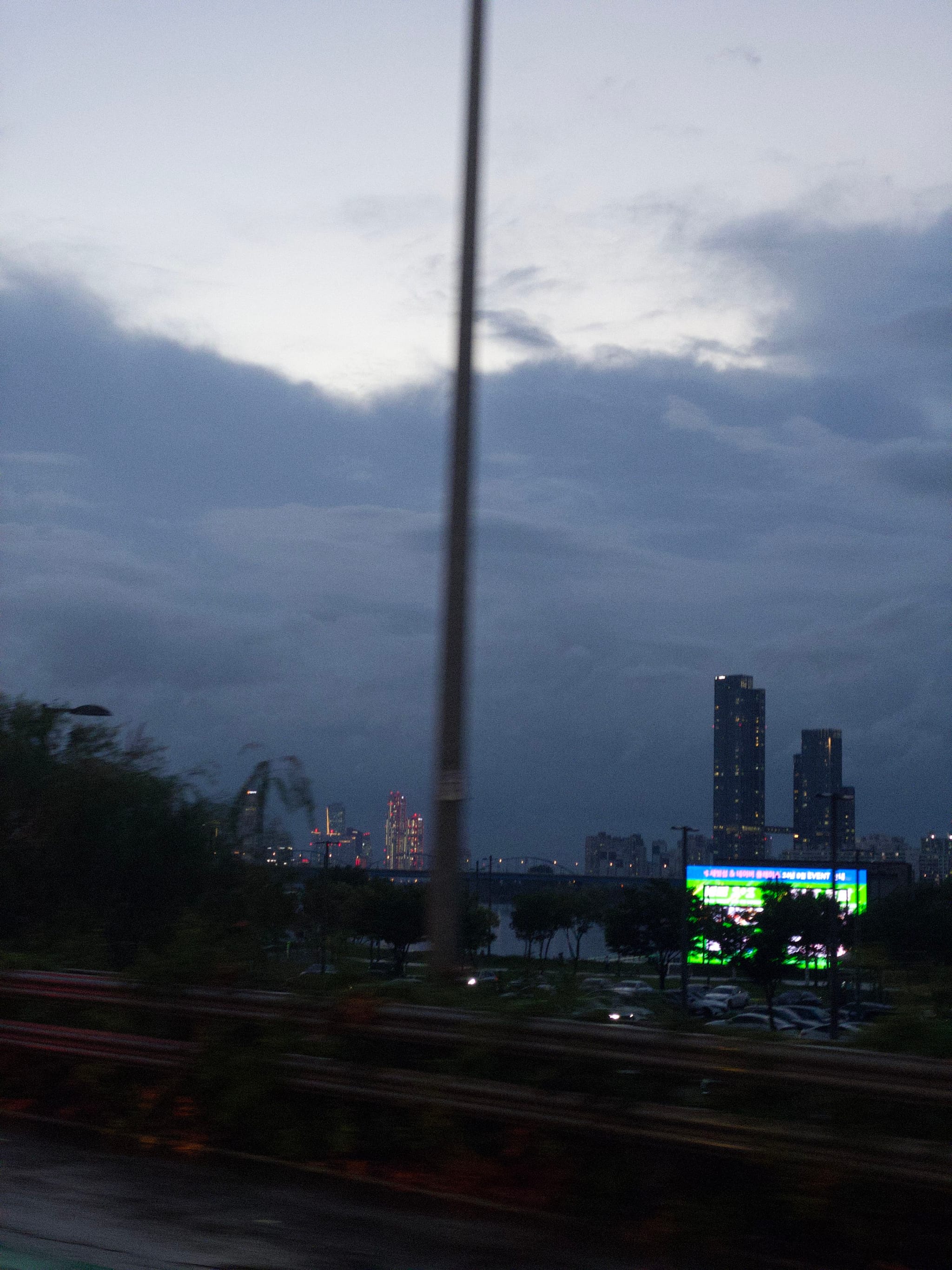 A cityscape at dusk with tall buildings in the background, a prominent streetlight in the foreground, and a large illuminated billboard