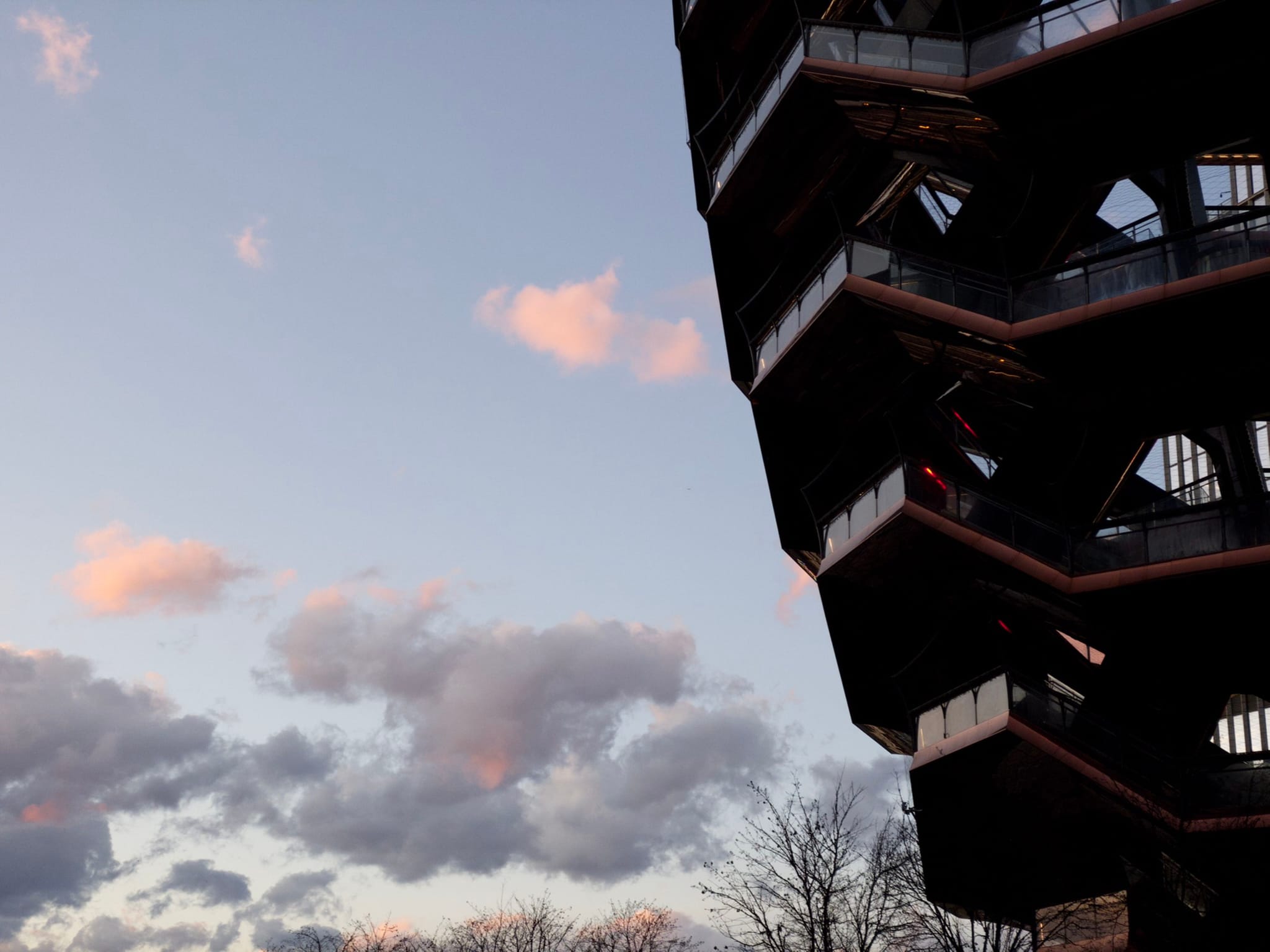 A modern, geometric building with multiple levels is silhouetted against a sky with scattered clouds, some tinged with pink