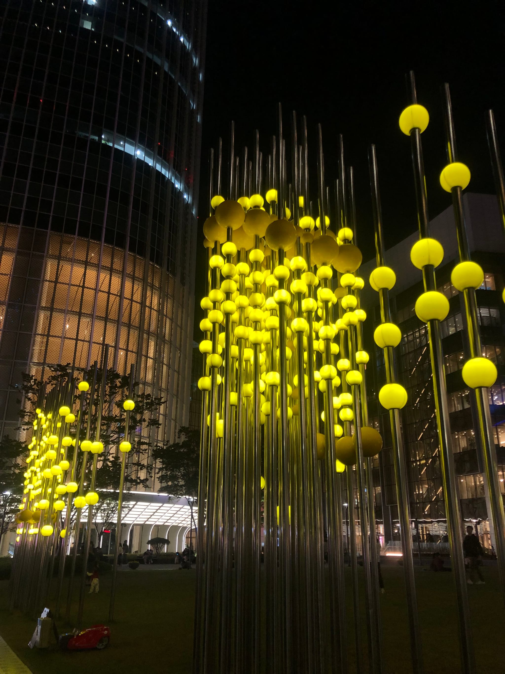 A nighttime scene featuring tall, illuminated yellow spheres on poles, set against a backdrop of modern buildings