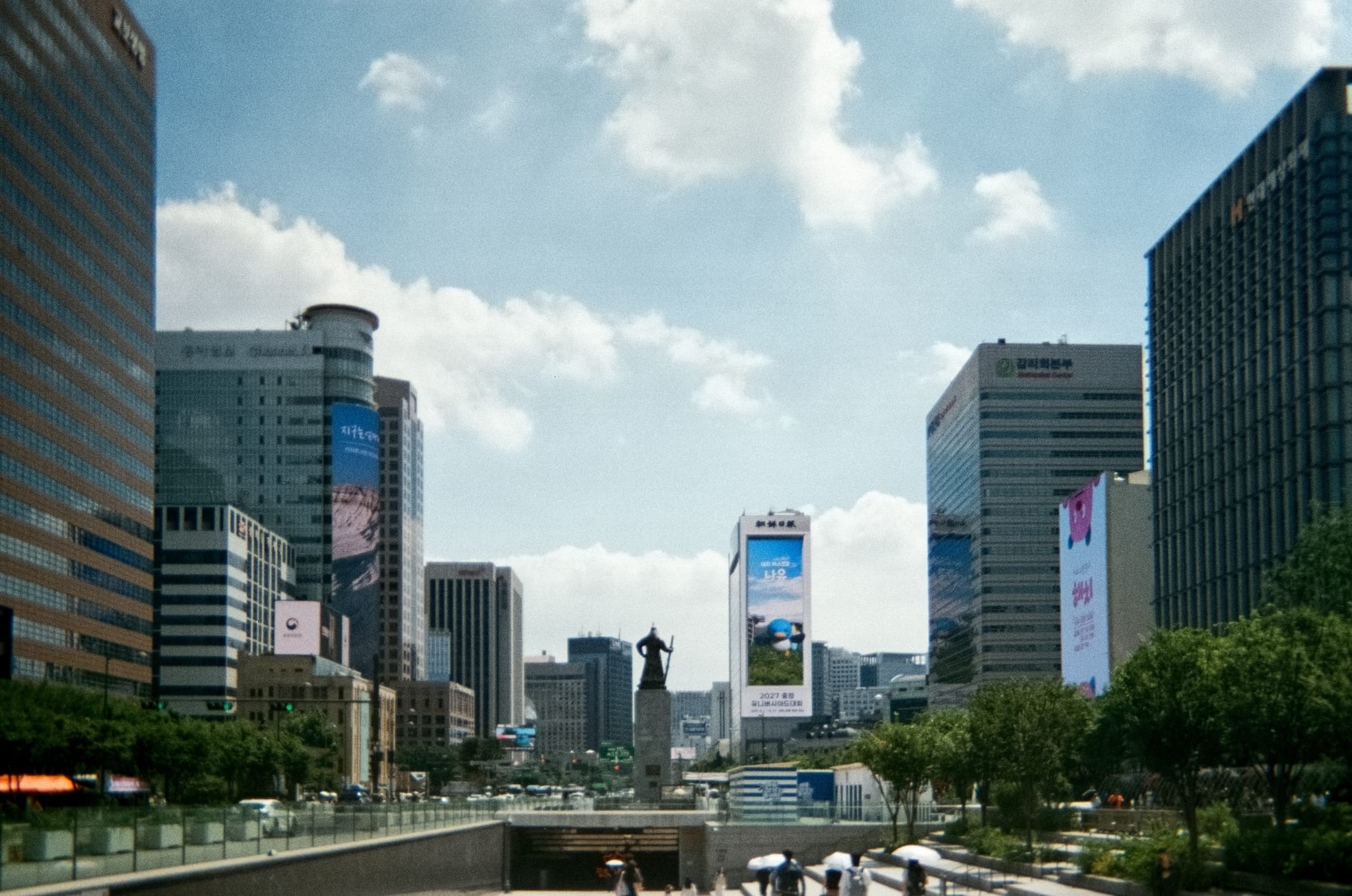 Wide boulevard in downtown Seoul lined with high-rise office buildings under scattered clouds