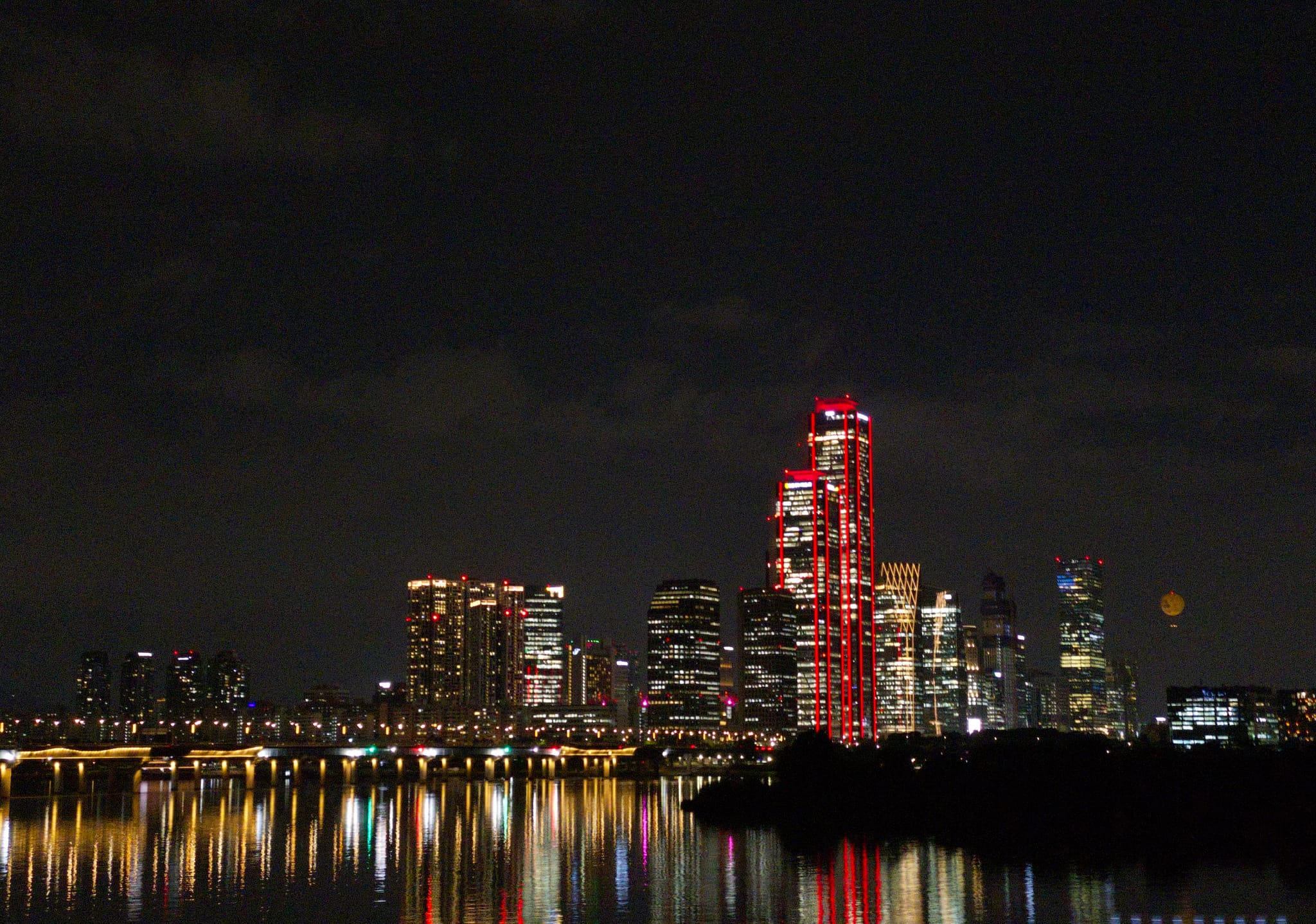 A city skyline at night with illuminated buildings, including one prominently lit in red, reflecting on a body of water