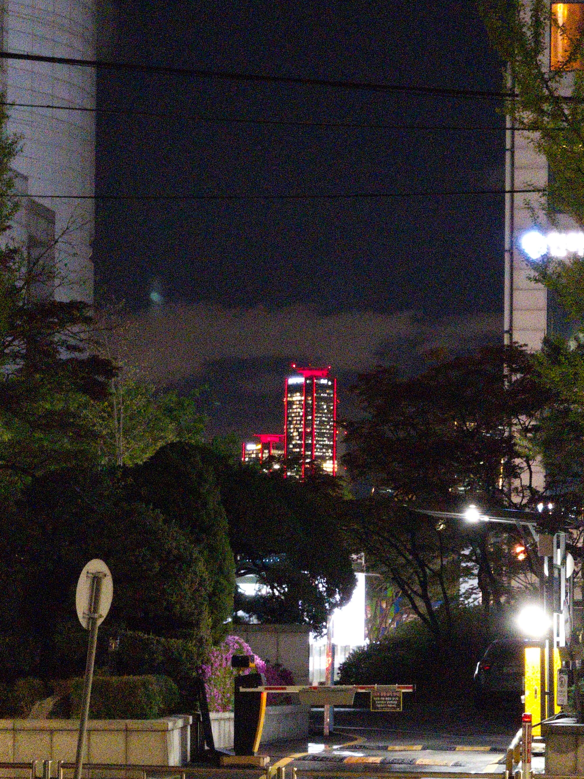 A nighttime urban scene with a distant illuminated building featuring red lights, framed by trees and structures in the foreground