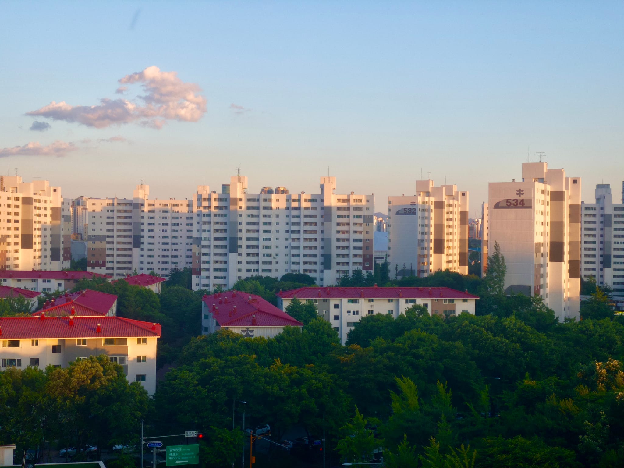 A cityscape featuring tall white apartment buildings in the background, with shorter buildings with red roofs and lush green trees in the foreground, under a clear blue sky with a few clouds