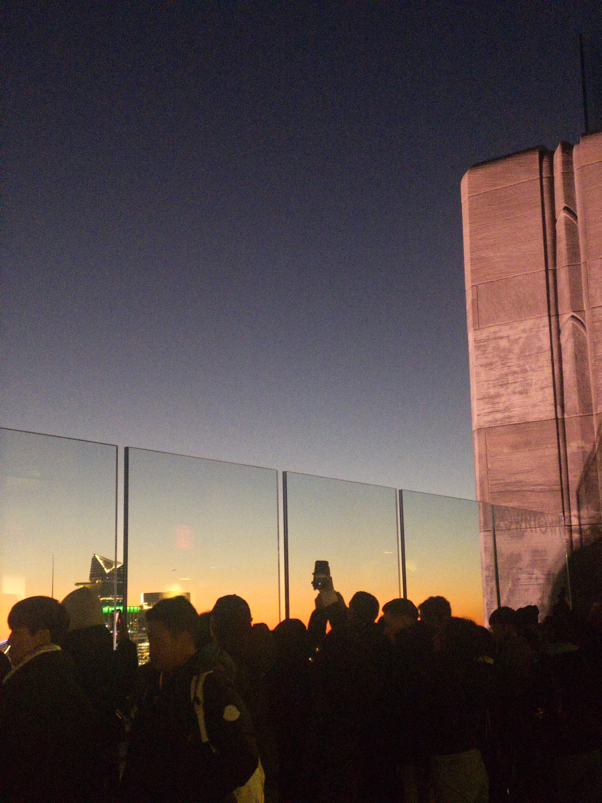 A crowd of people stands on a rooftop observation deck at sunset, with a clear sky and a large building structure on the right