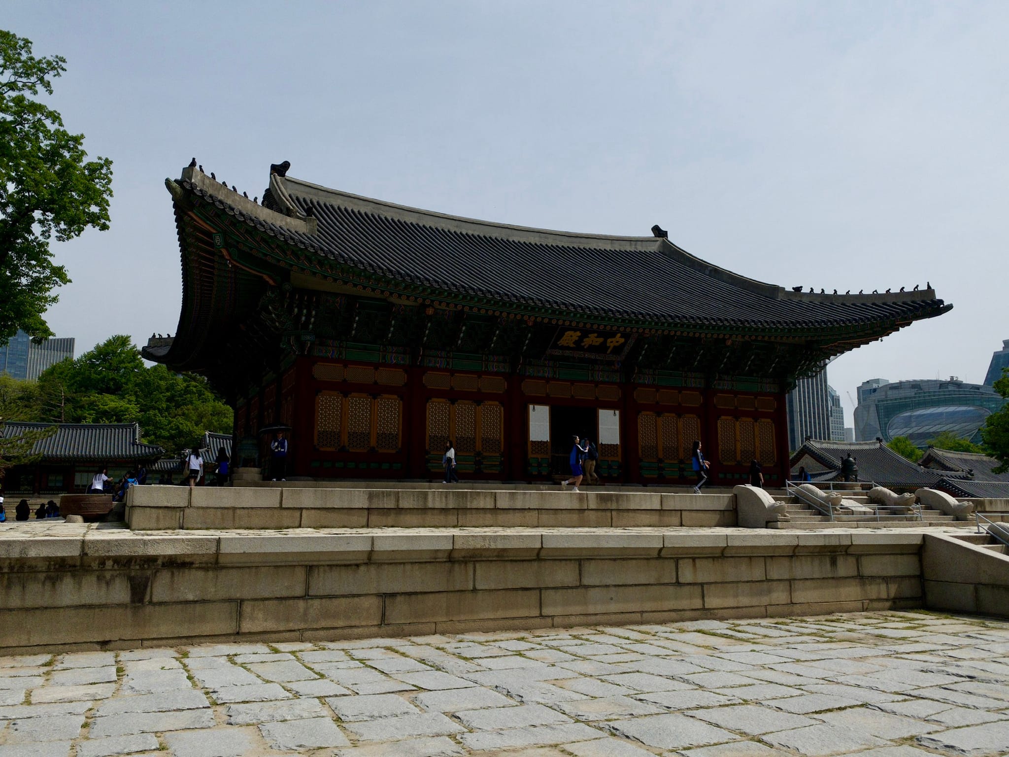 A traditional Korean building with a curved roof, set on a stone platform, surrounded by trees and modern buildings in the background