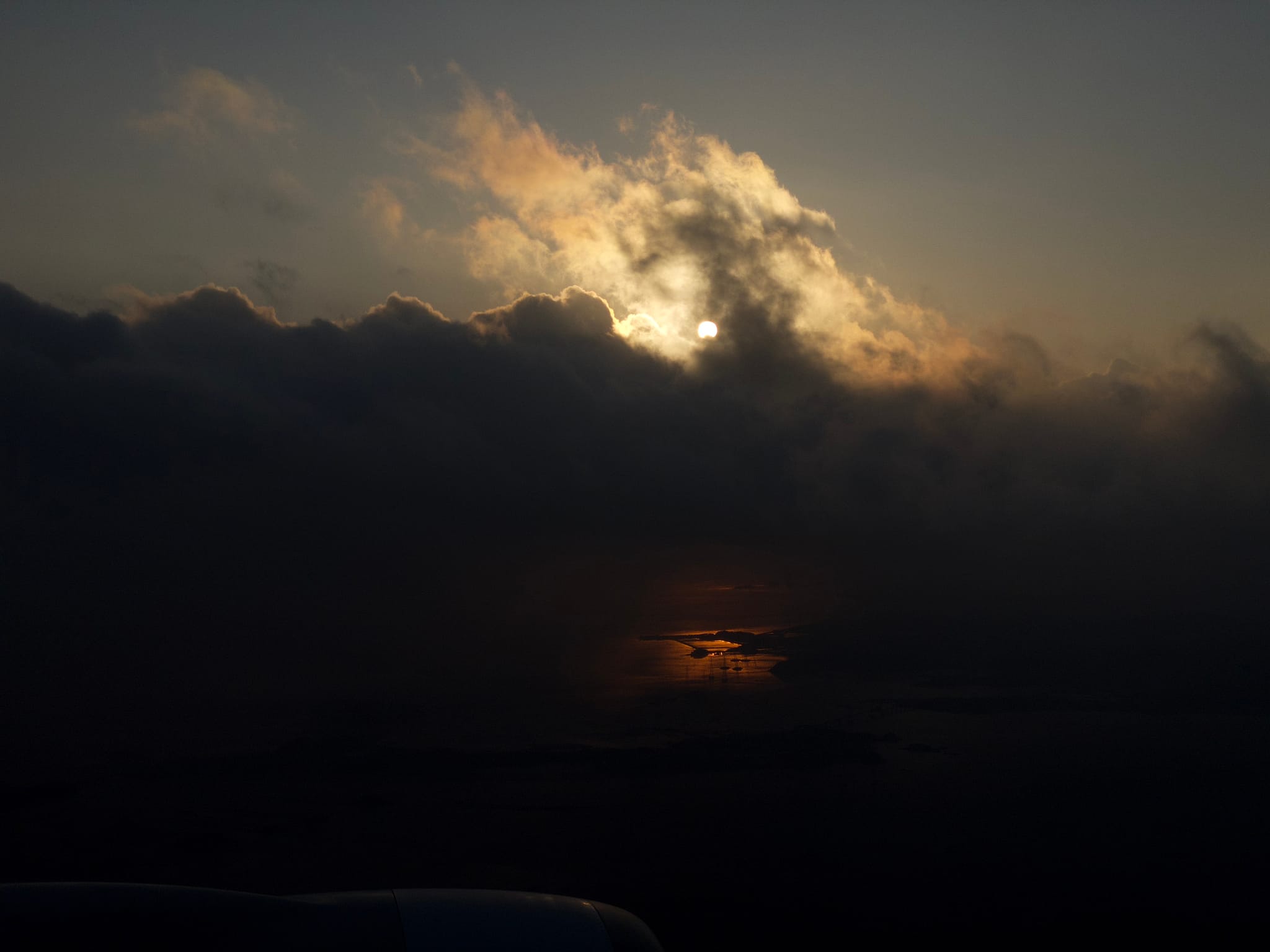 A dramatic sky with the sun partially obscured by clouds, casting a warm reflection on a dark landscape below