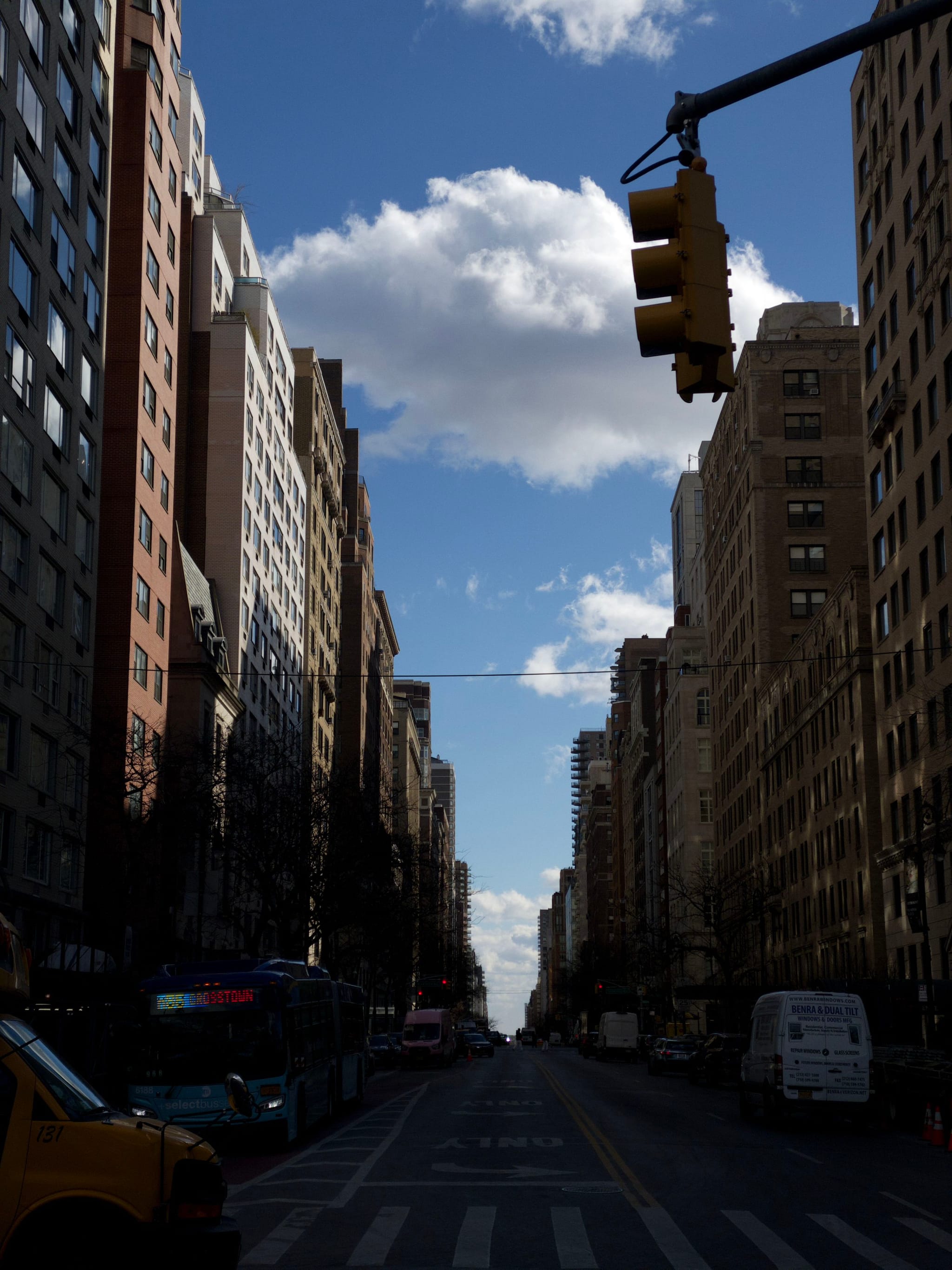 A city street lined with tall buildings under a blue sky with clouds, featuring a traffic light and vehicles