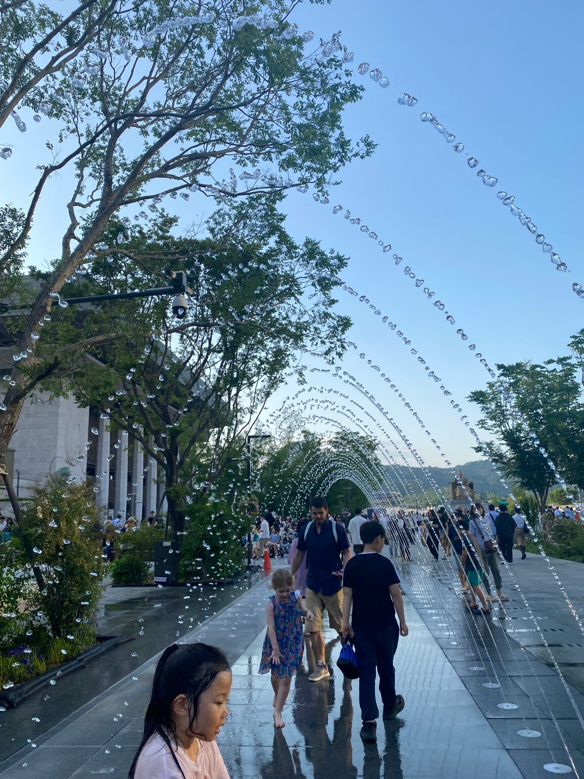 A park scene with people walking under arched water fountains, surrounded by trees and greenery