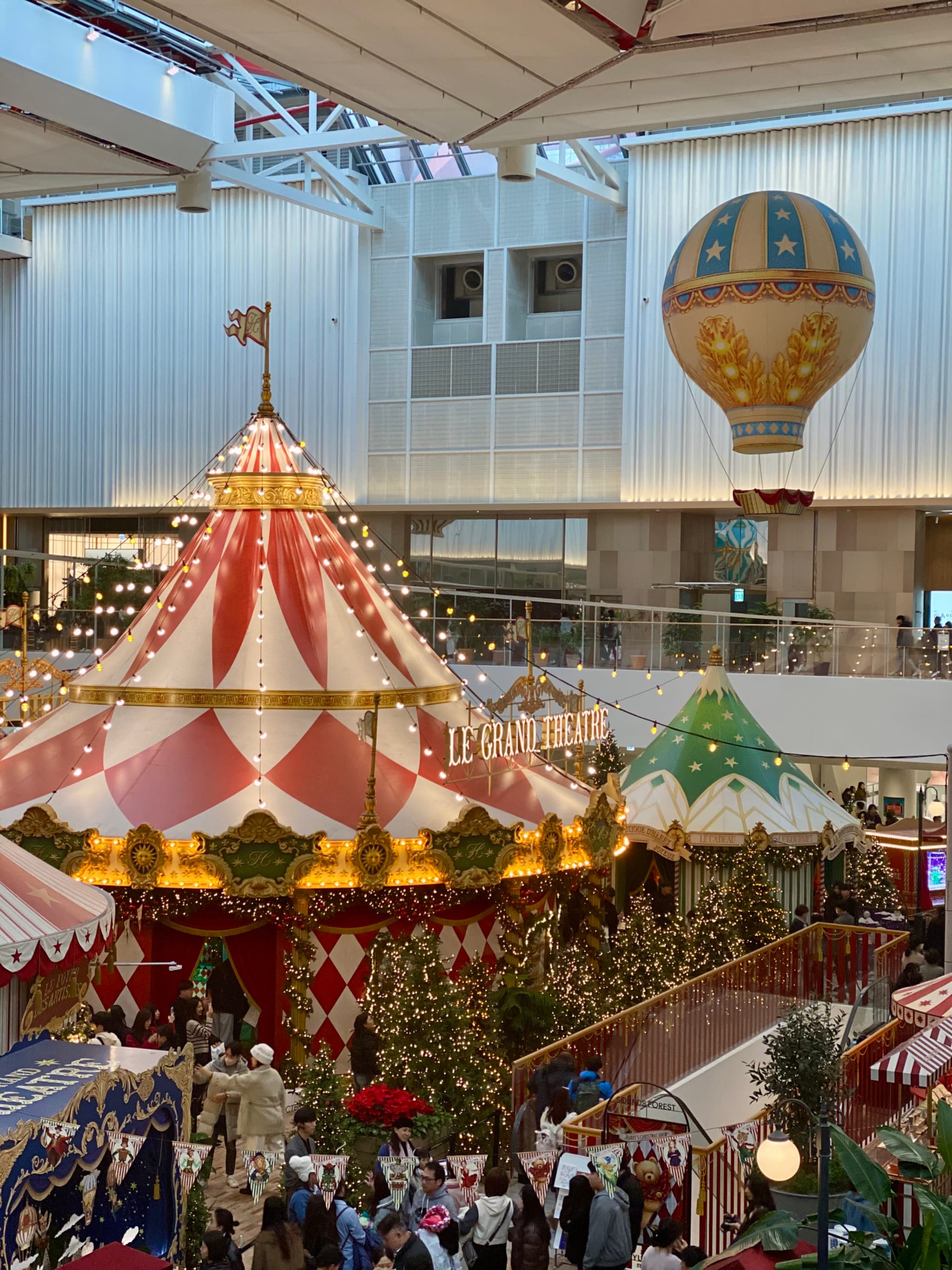 A festive indoor scene with colorful circus tents, string lights, and a hot air balloon decoration, surrounded by Christmas trees and bustling with people