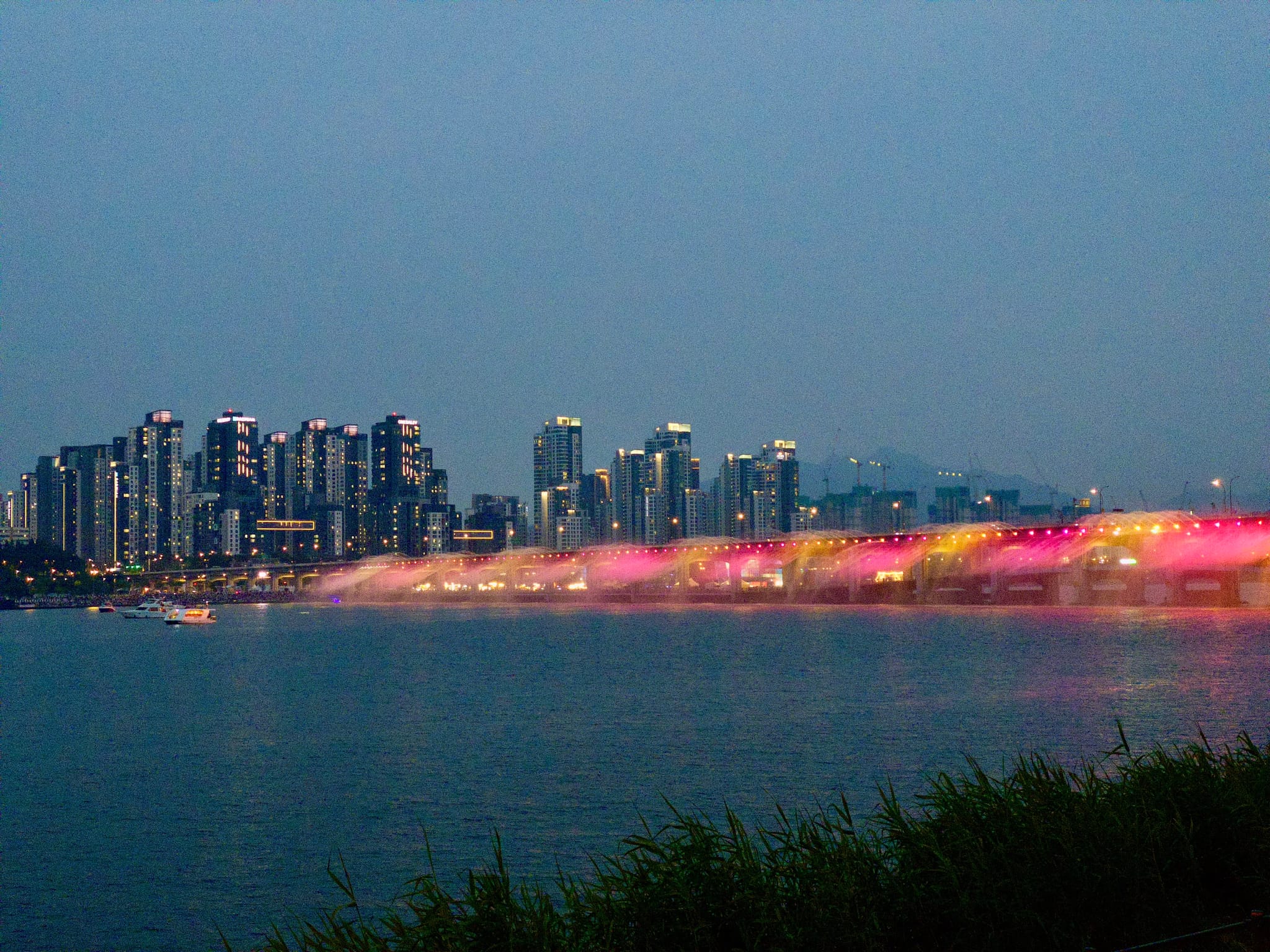 A city skyline at dusk with illuminated fountains creating colorful light illuminated fountains over a river, and grassy foreground