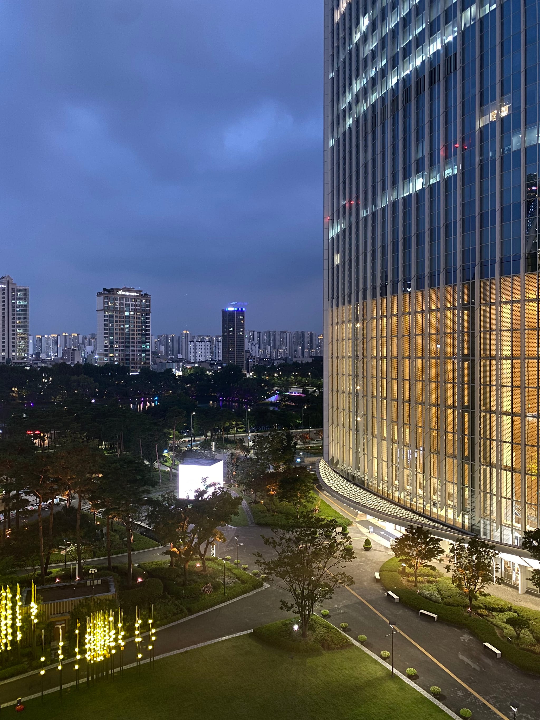 A cityscape at dusk with a tall, illuminated building on the right, surrounded by trees and roads, and other buildings visible in the background under a cloudy sky