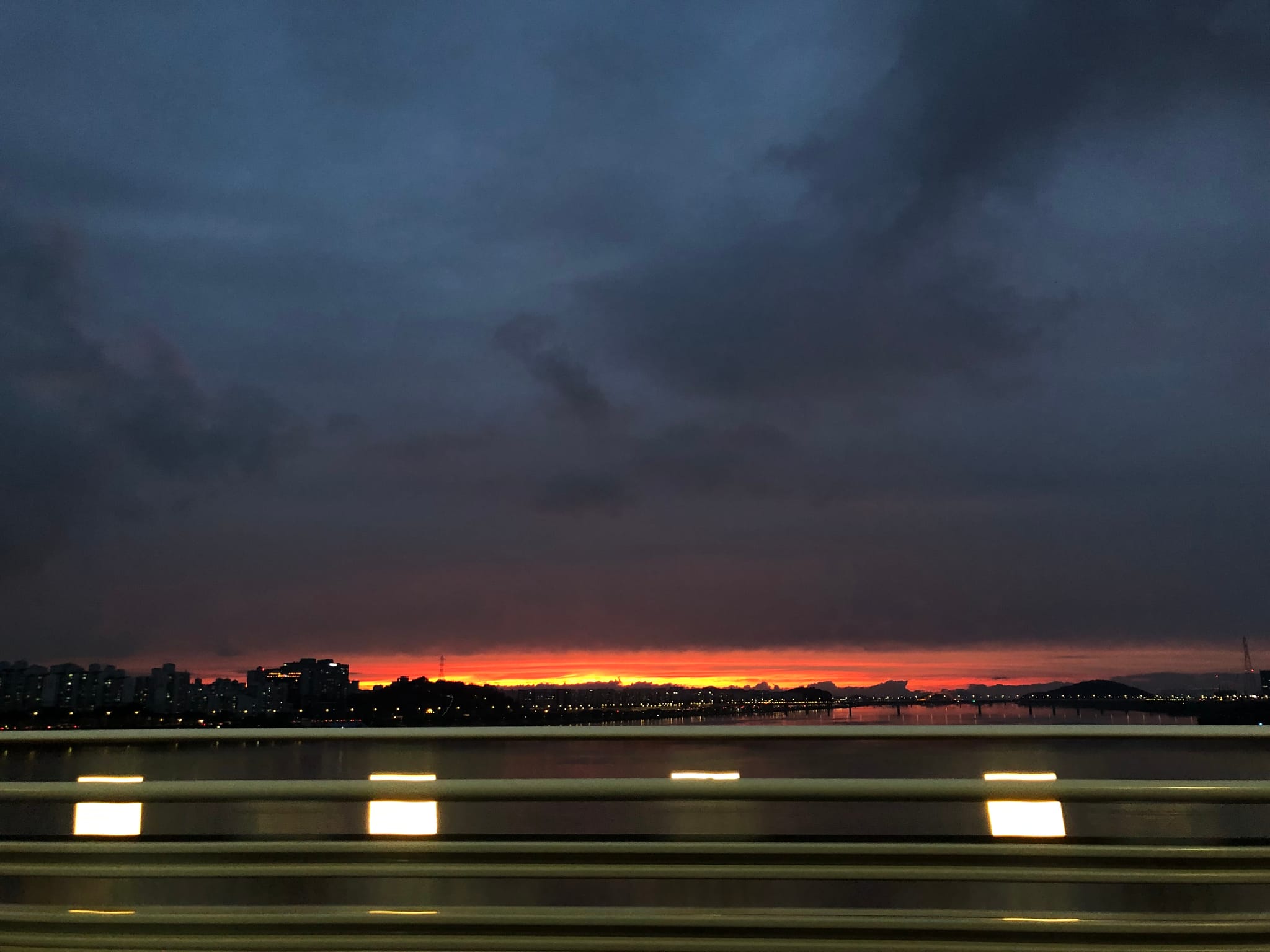 A dark, cloudy sky with a vibrant orange and yellow sunset on the horizon, viewed from behind a blurred railing or barrier