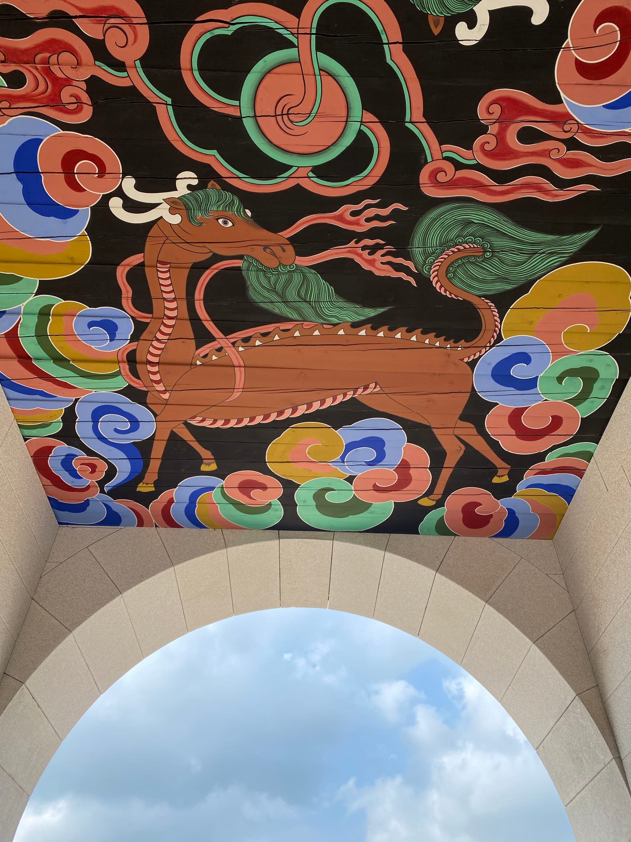 A colorful, ornate ceiling mural featuring a stylized dragon surrounded by swirling clouds and patterns, viewed through an archway with a glimpse of the sky
