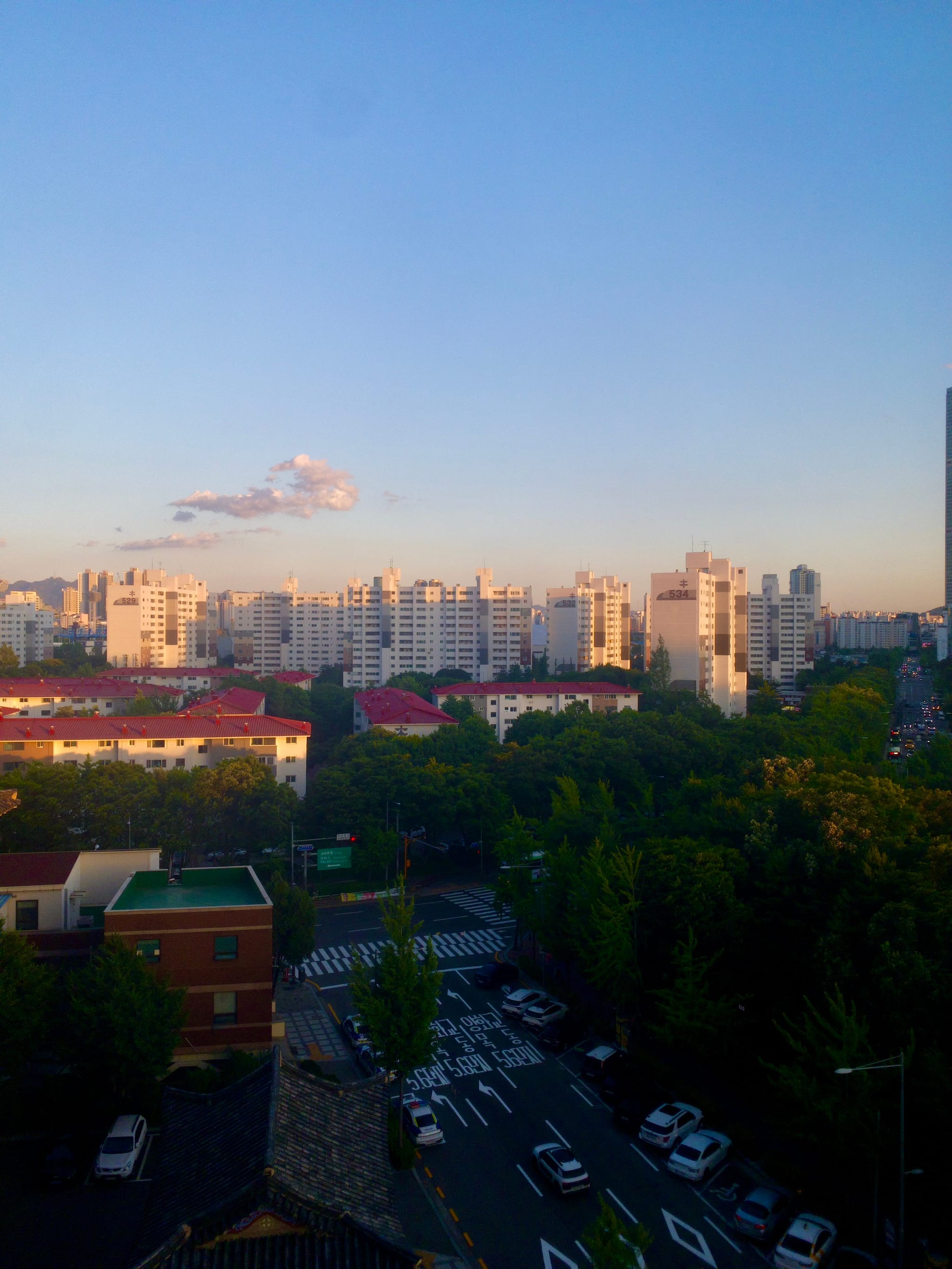 A cityscape at sunset with tall buildings in the background, a few clouds in the sky, and a tree-lined street with cars in the foreground
