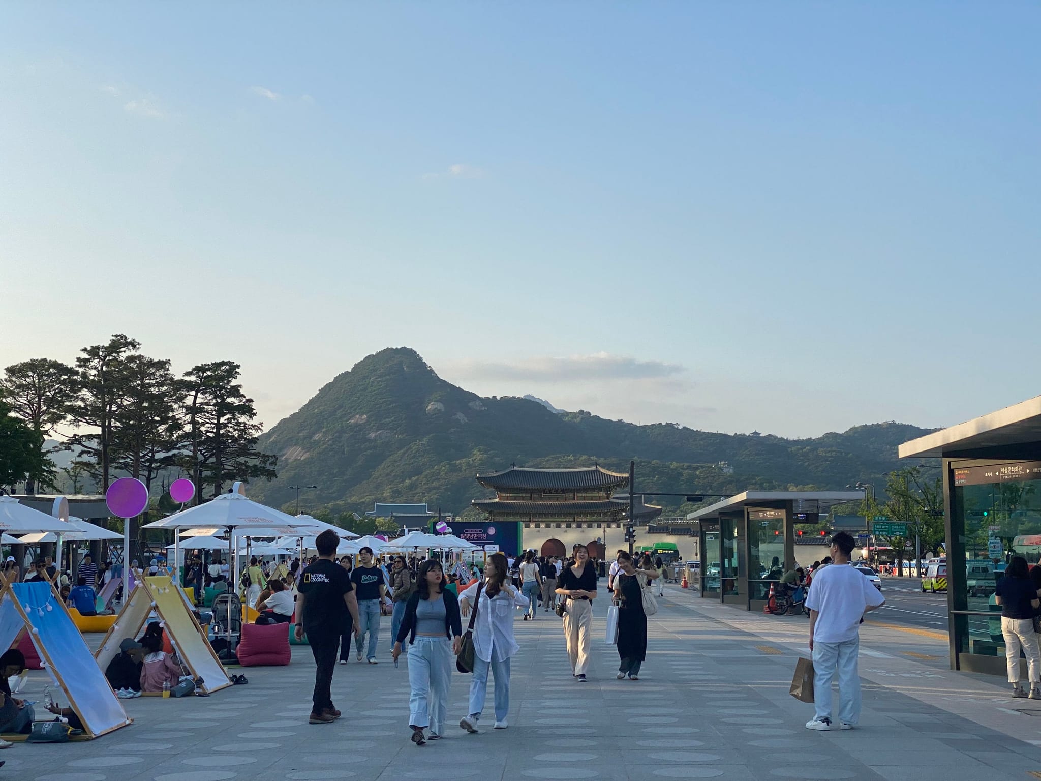 A bustling outdoor market with people walking along a wide pathway, surrounded by tents and stalls. A mountain and clear sky are visible in the background