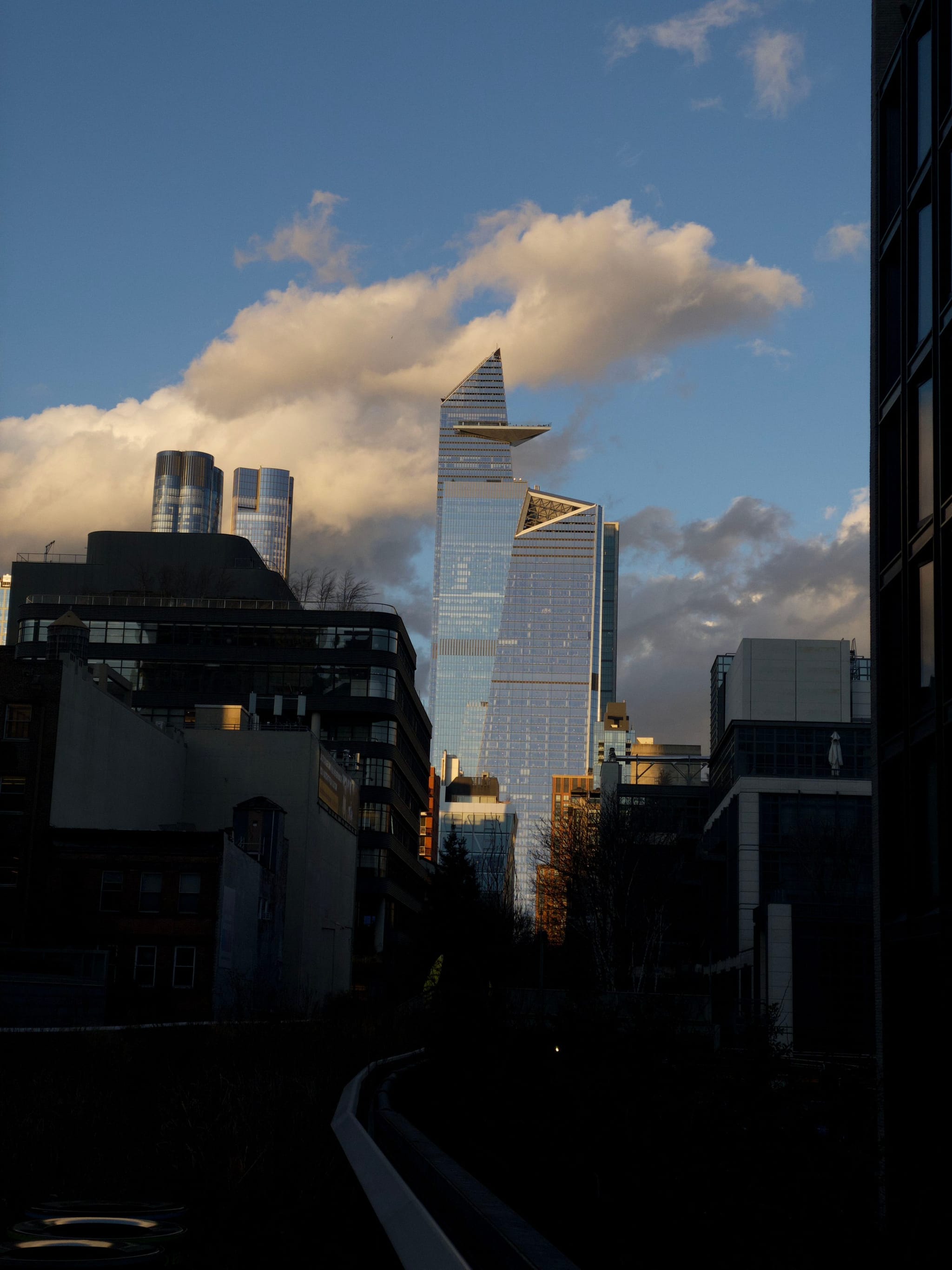 A cityscape featuring modern skyscrapers with reflective glass facades under a partly cloudy sky, with sunlight illuminating the buildings