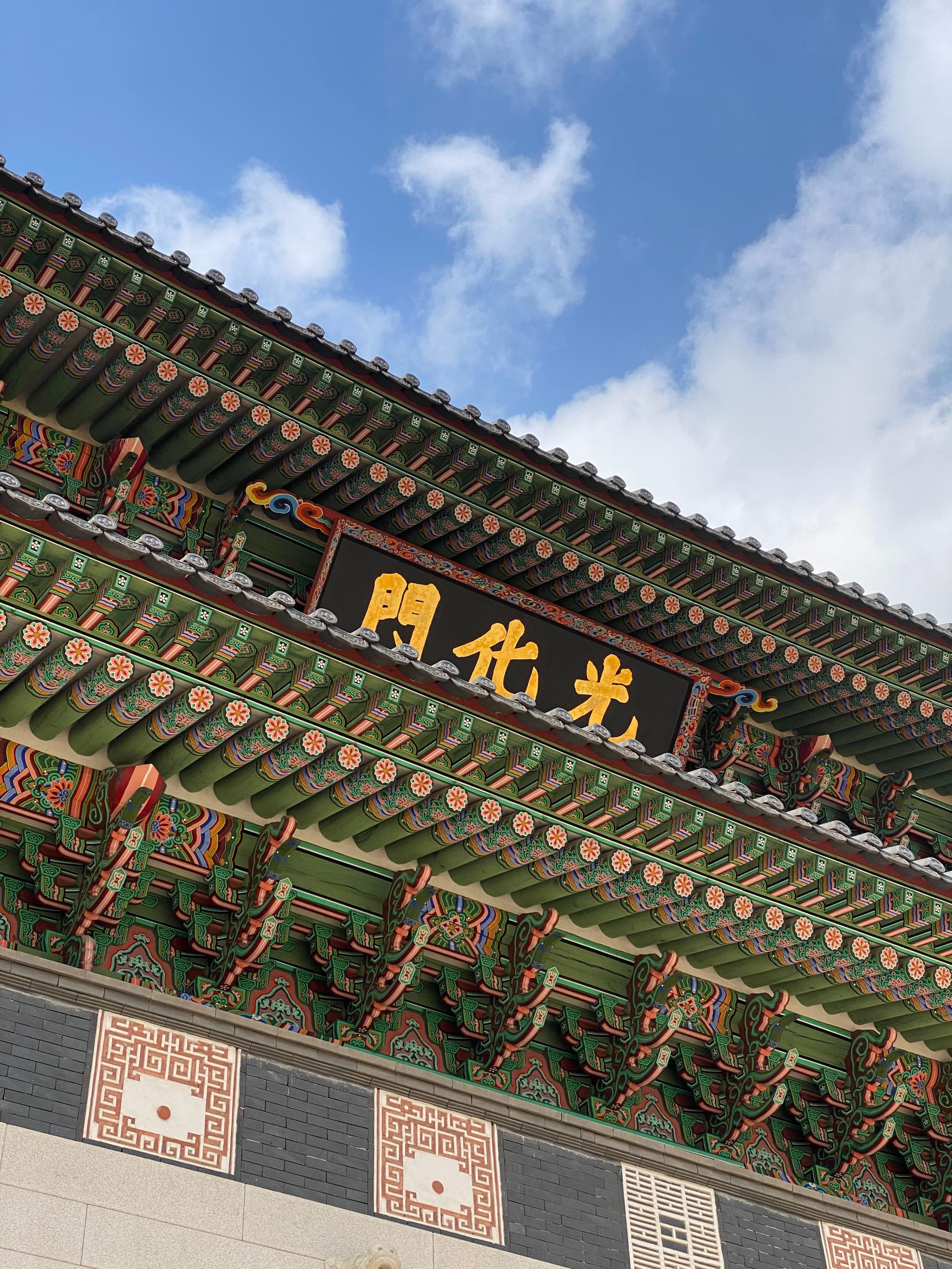 A traditional Korean architectural structure with ornate, colorful eaves and a sign with Korean characters, set against a blue sky with clouds