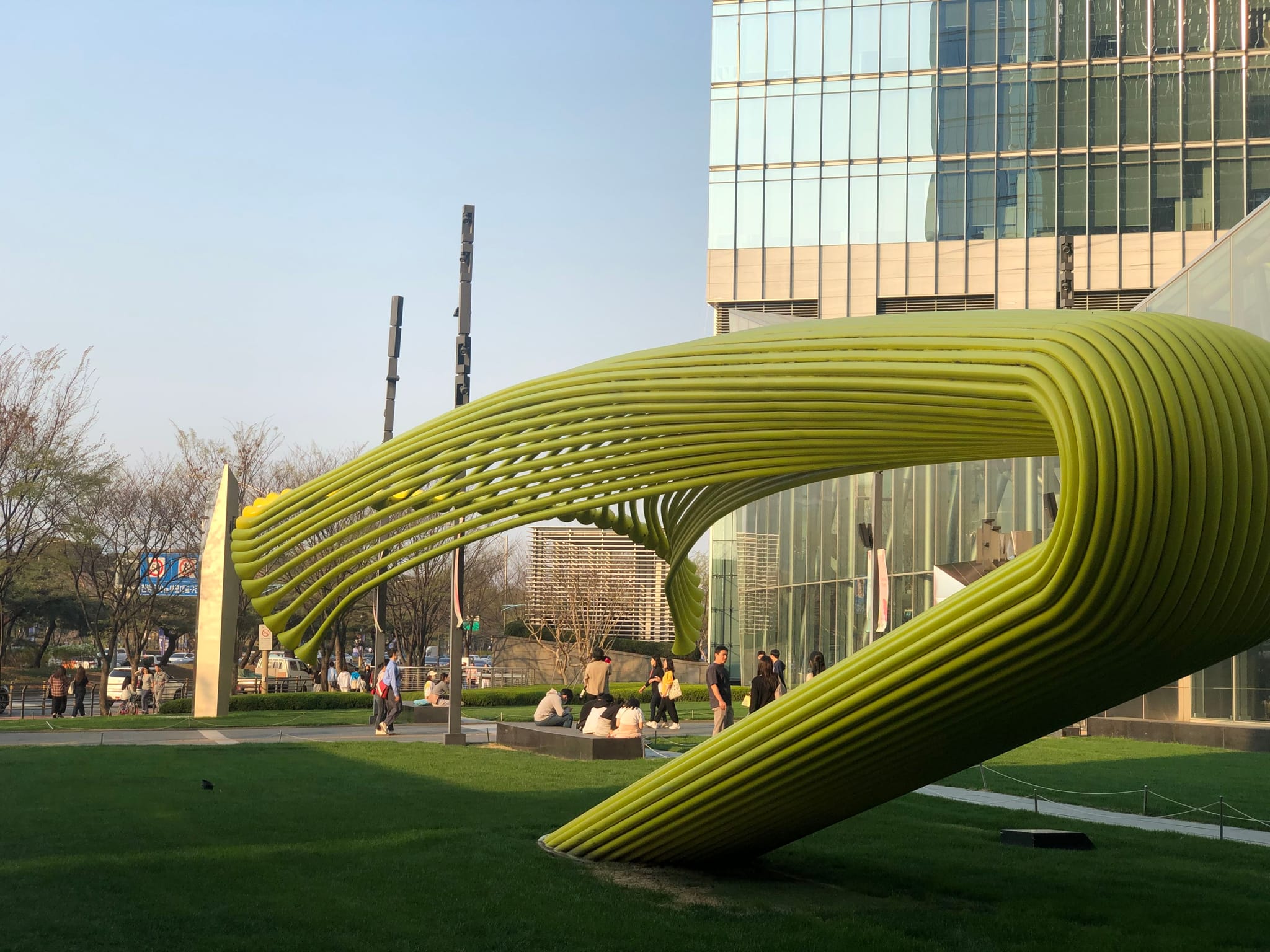 A large, modern yellow sculpture with a curved, ribbed design is situated on a grassy area near a glass building. People are walking nearby, and trees are visible in the background