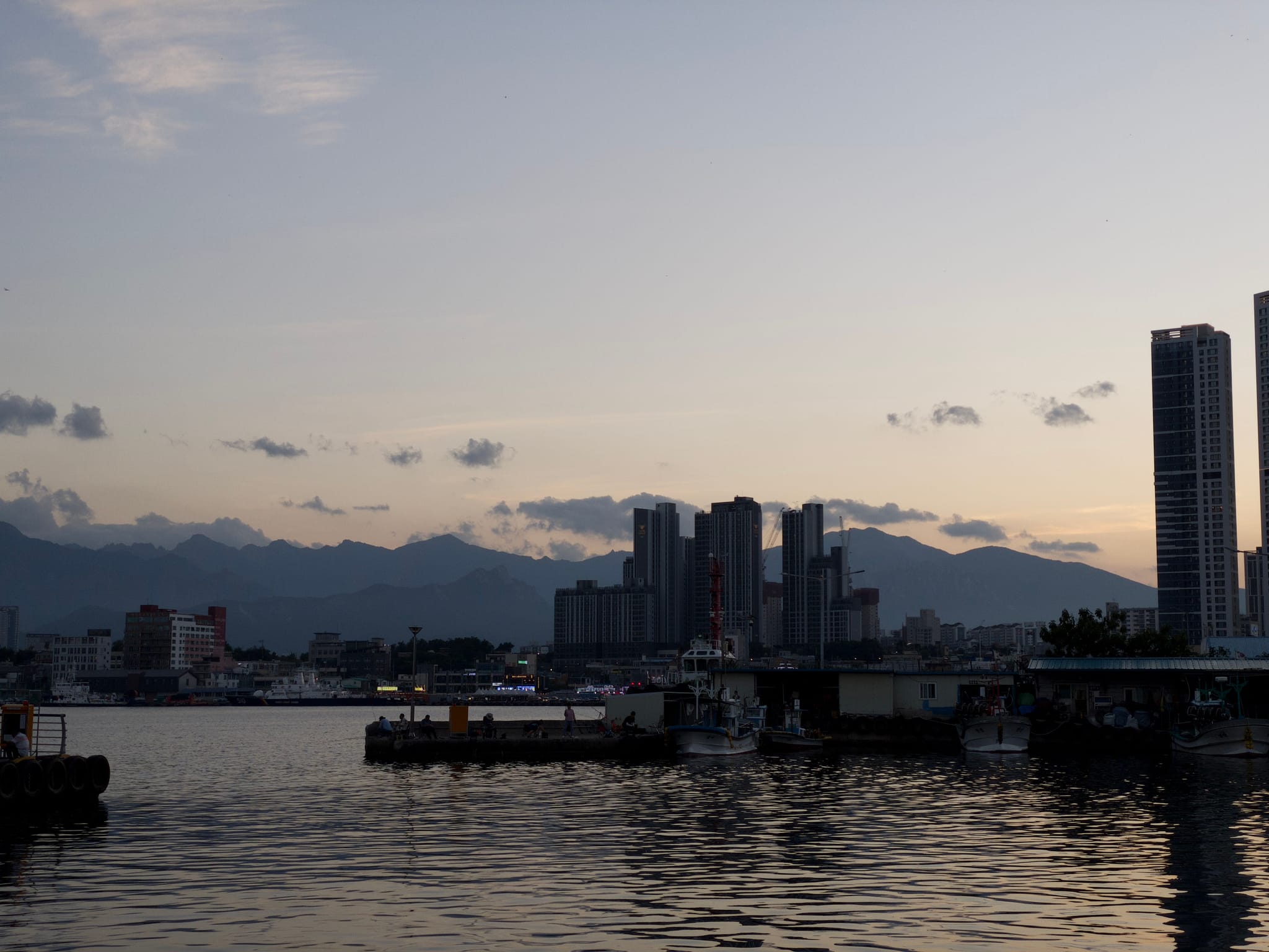 A city skyline at dusk with tall buildings silhouetted against a mountain backdrop and a body of water in the foreground