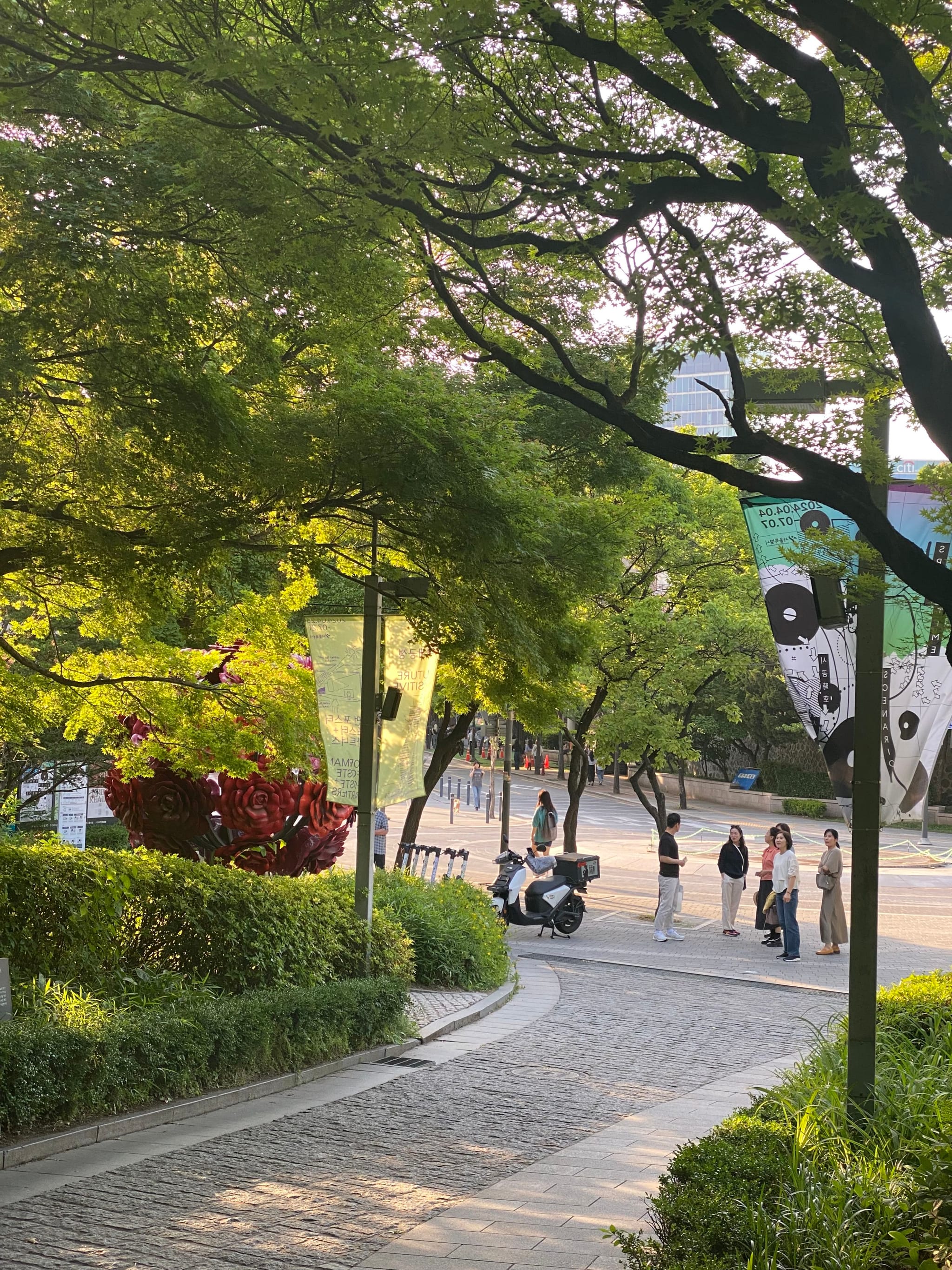 A sunlit park scene with a cobblestone path, lush green trees, and people walking. Banners hang from poles, and a scooter is parked nearby
