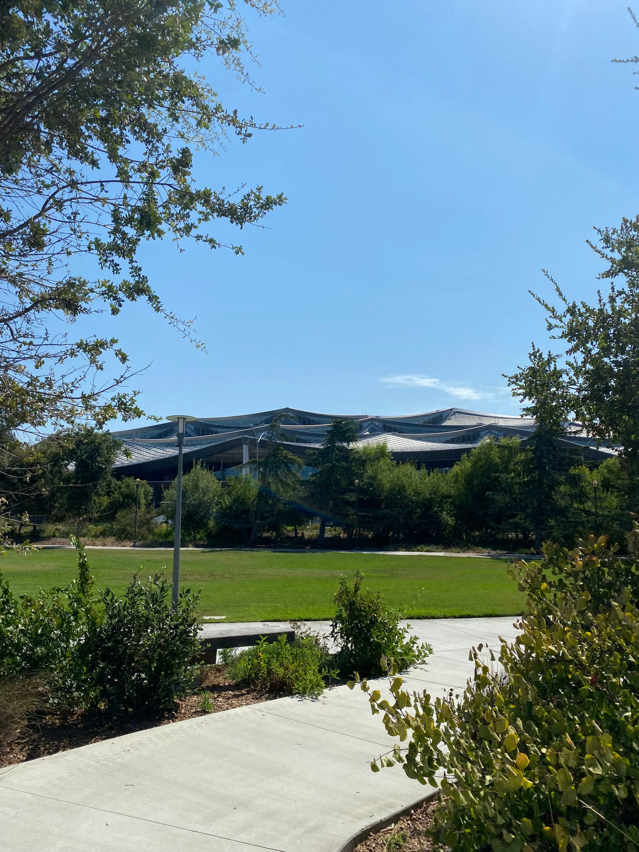 A modern building with a wavy roof is visible in the background, surrounded by trees and greenery. A paved pathway leads through a grassy area in the foreground under a clear blue sky