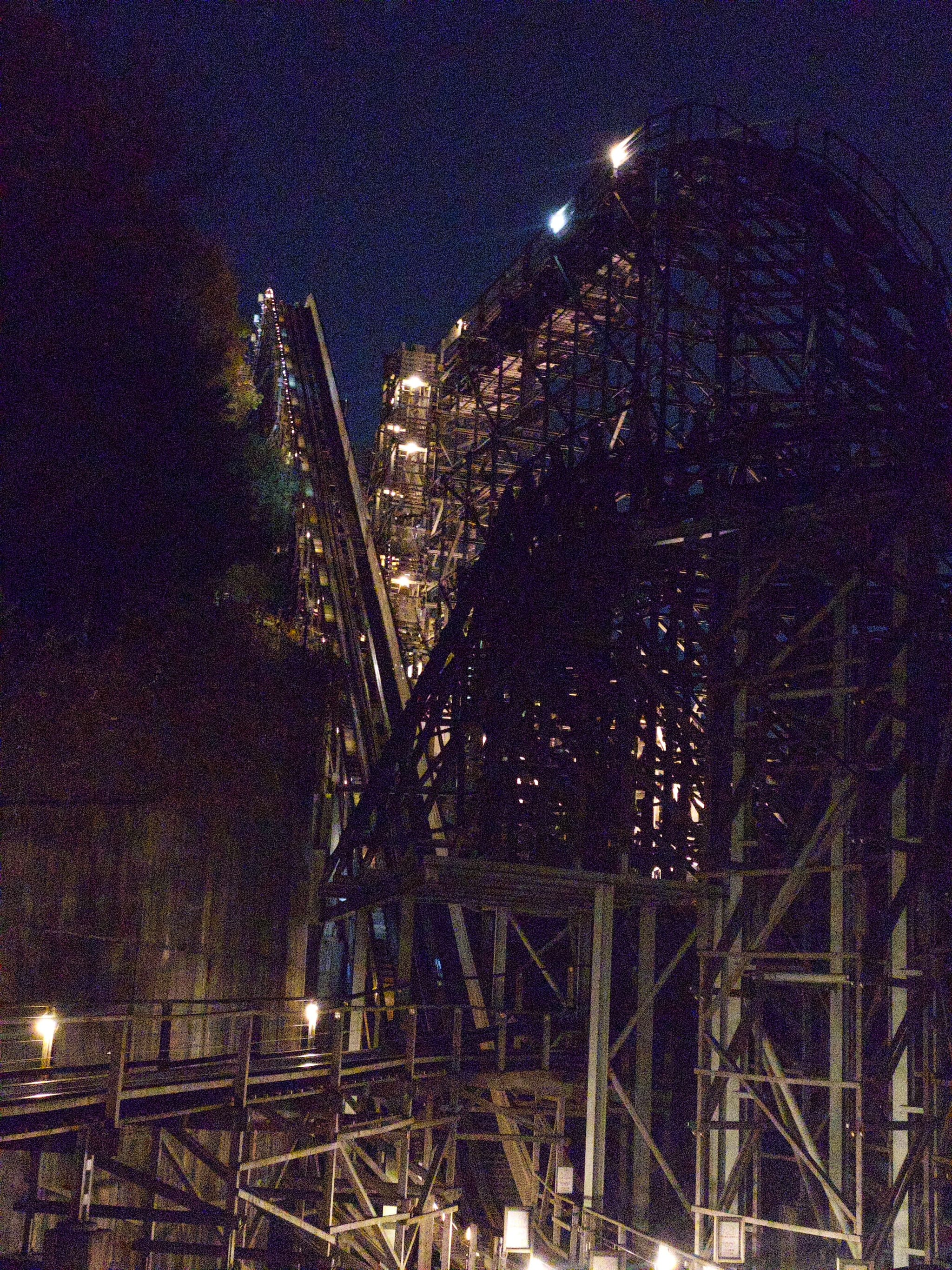 A wooden roller coaster structure is illuminated at night, with visible tracks and support beams