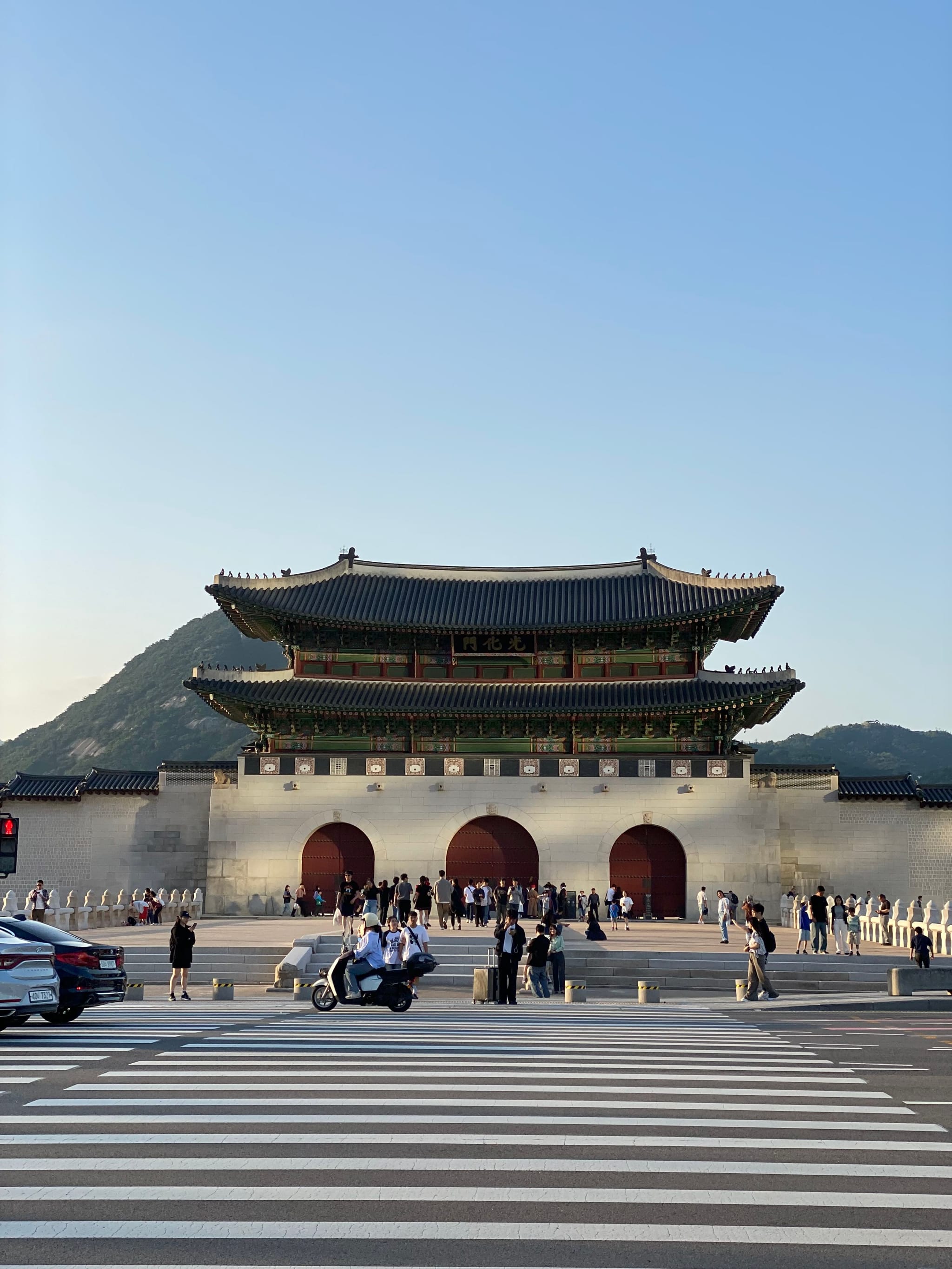 A traditional Korean gate with three arched entrances, set against a backdrop of mountains and a clear sky, with people and vehicles in the foreground