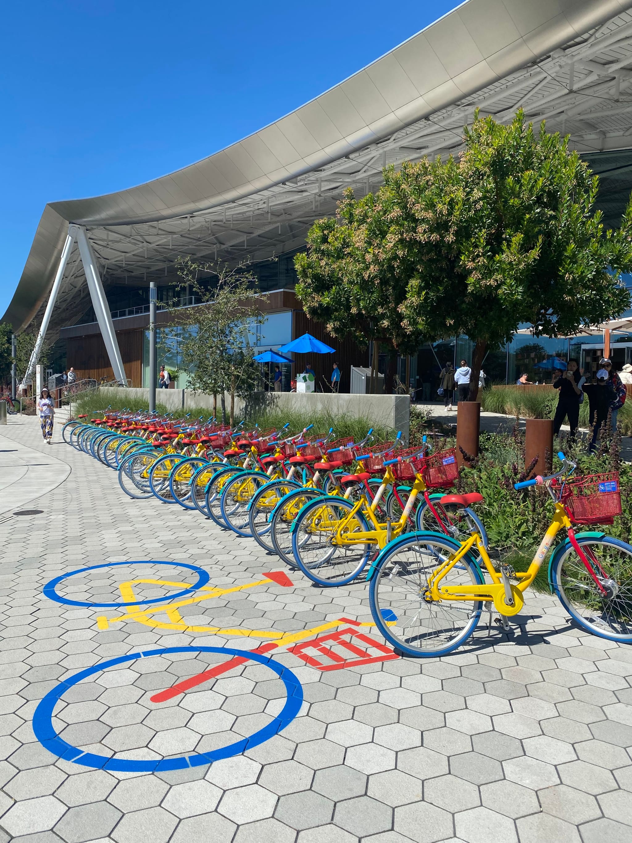 A row of colorful bicycles is lined up on a hexagonal-tiled pathway, with a modern building and trees in the background under a clear blue sky