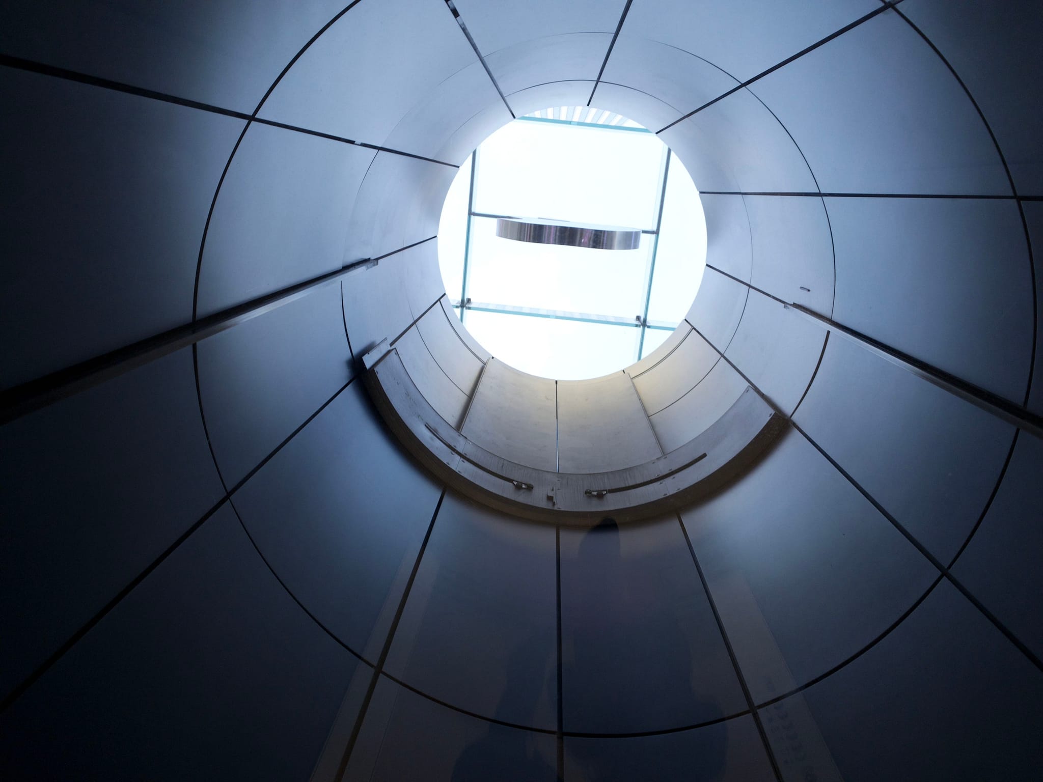 A view looking up through a circular, tunnel-like structure with segmented panels, leading to an opening at the top that lets in light