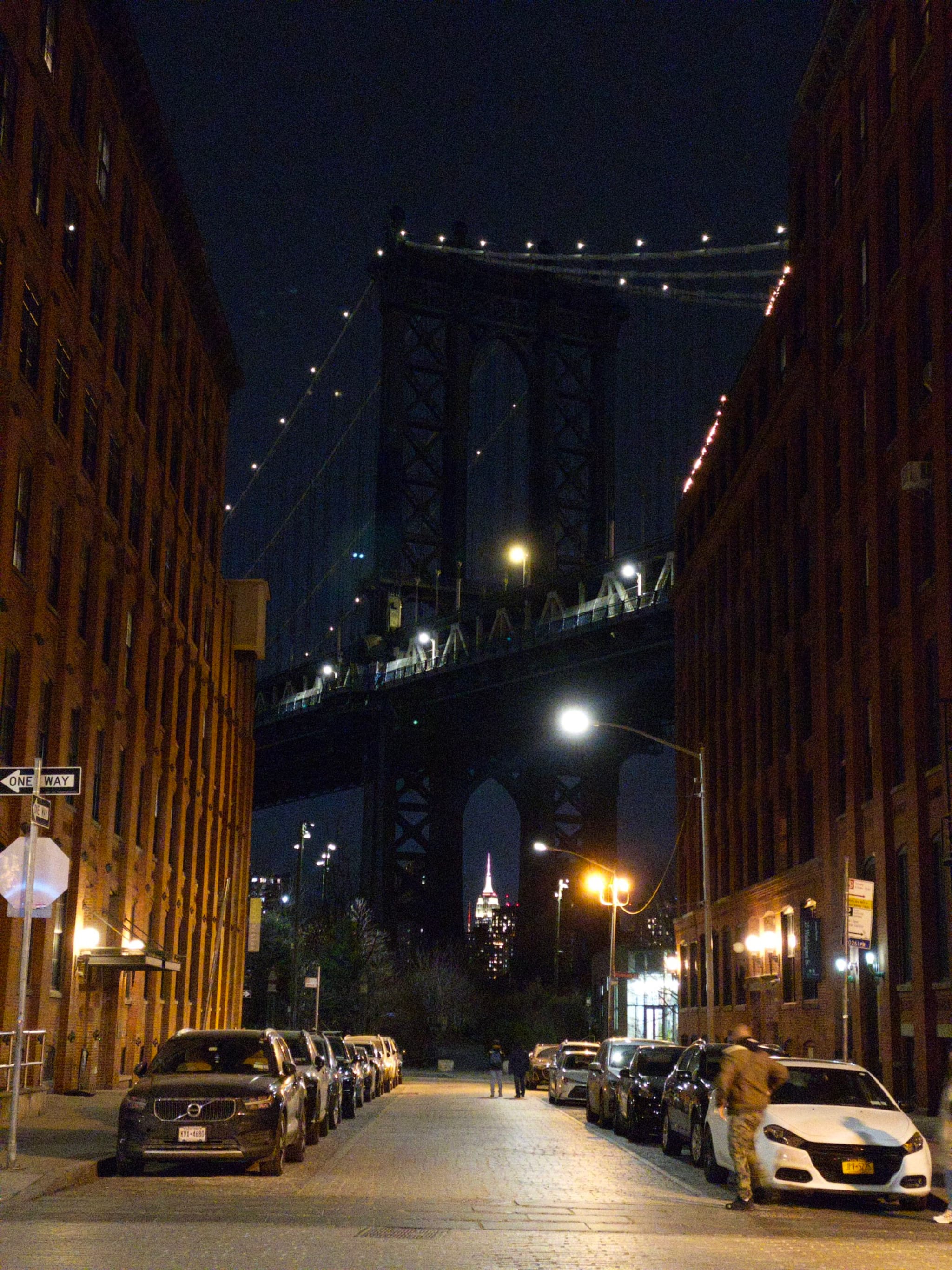 A nighttime view of a city street with parked cars on both sides, leading to a large bridge in the background, illuminated by streetlights