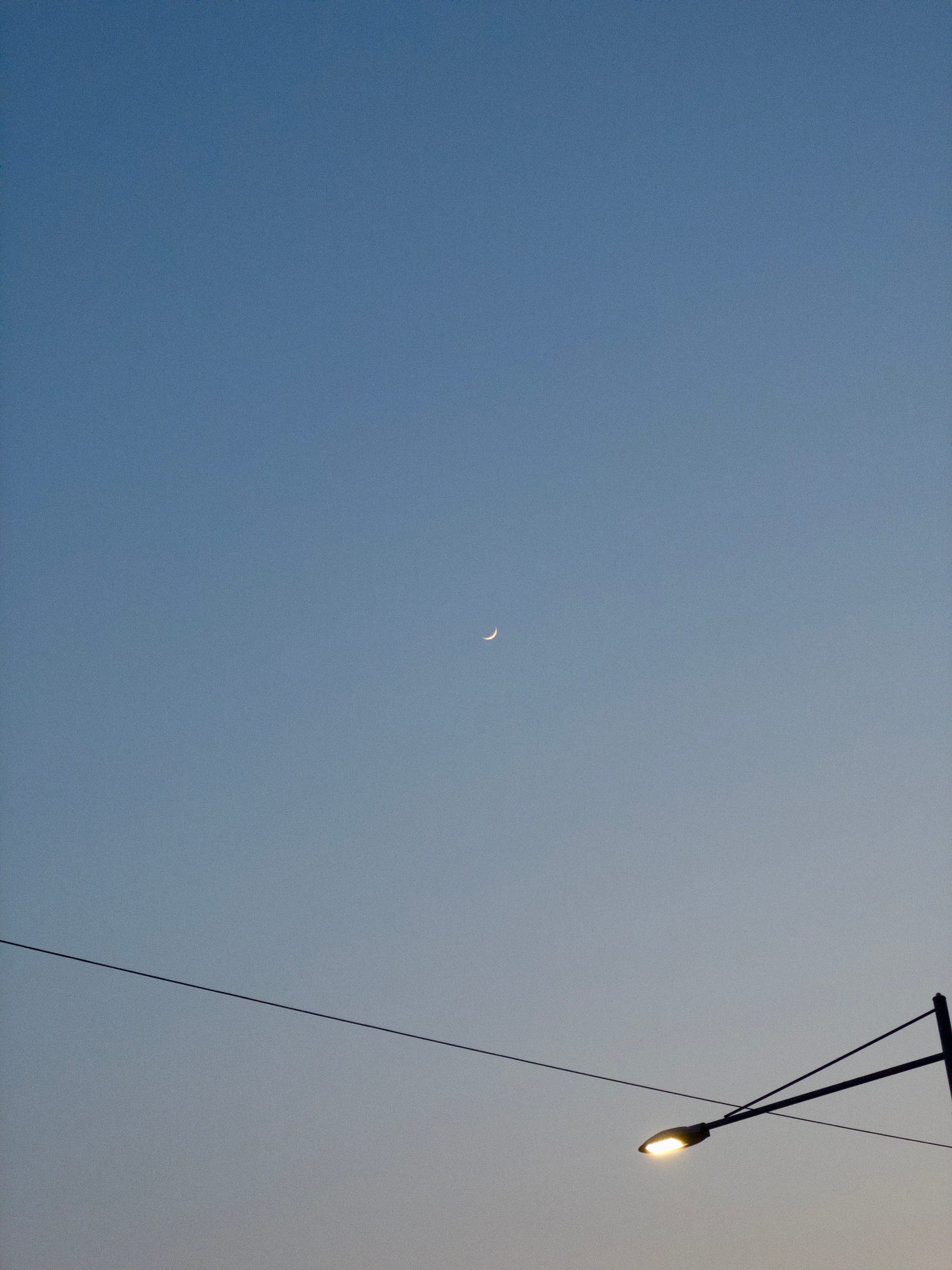 A crescent moon in a clear evening sky with a streetlight and power lines in the foreground