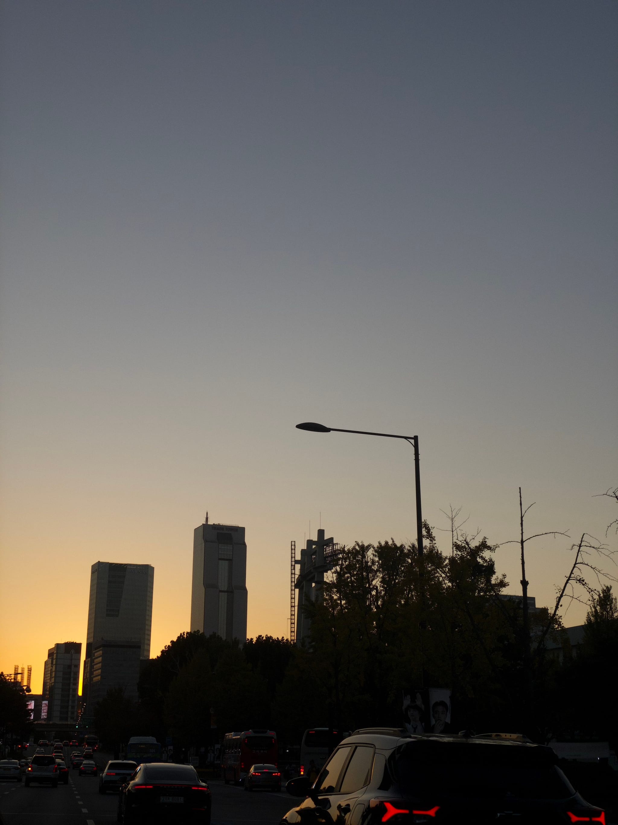 A cityscape at dusk with tall buildings silhouetted against a gradient sky, cars on the road, and a streetlamp in the foreground