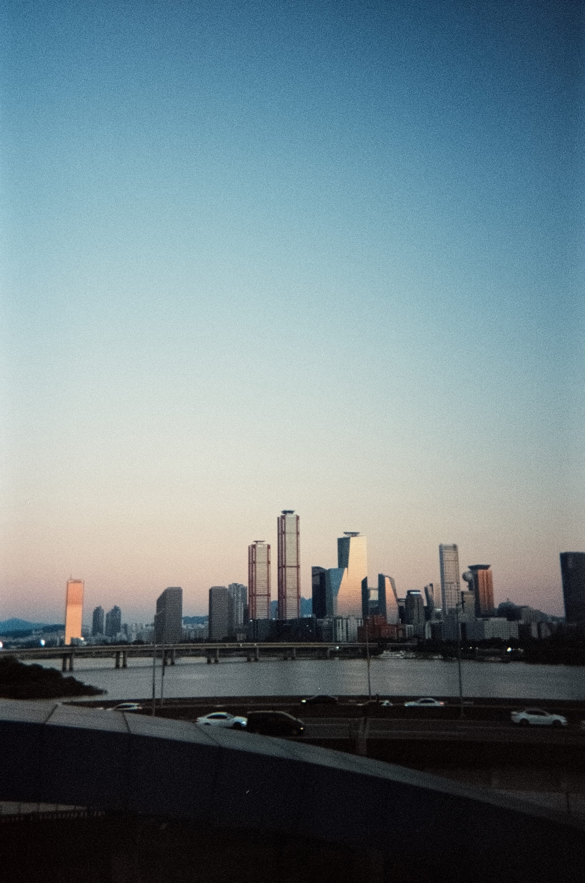 Distant urban skyline with high-rise towers across a river at dusk, viewed from a bridge railing under a clear gradient sky
