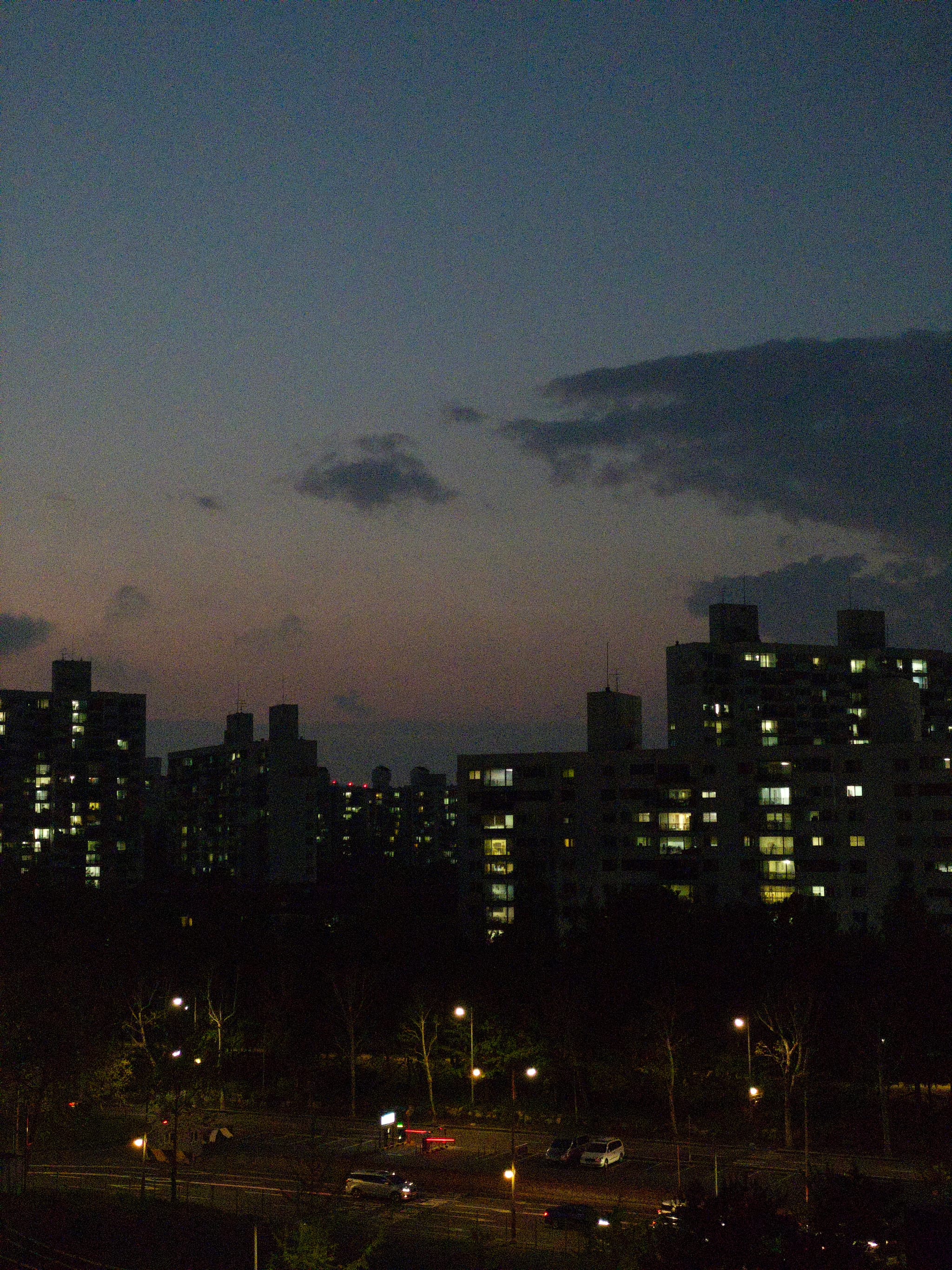 A cityscape at dusk with illuminated buildings and a darkening sky, featuring a few clouds and streetlights below