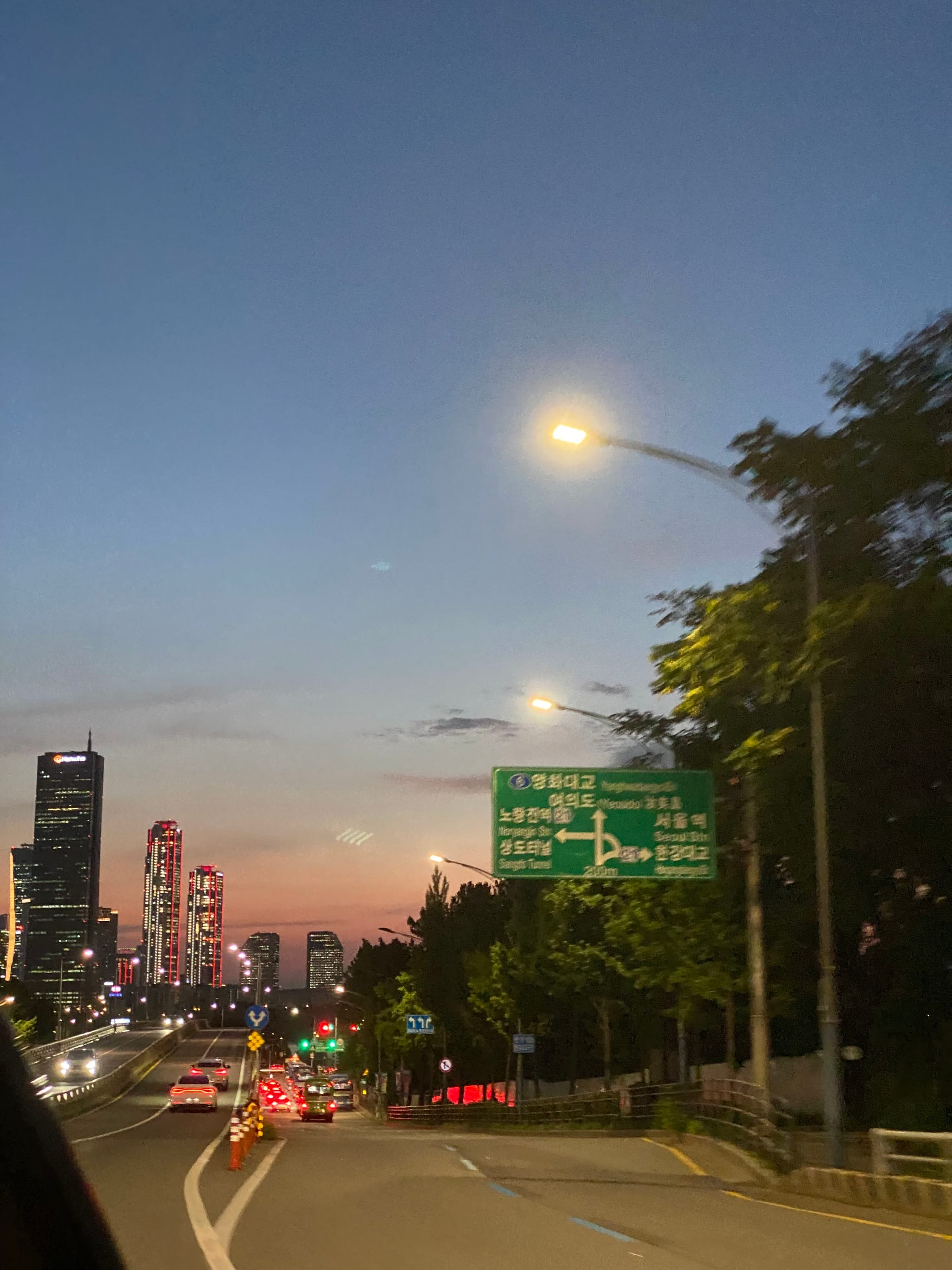 A cityscape at dusk with a road leading towards tall buildings, streetlights illuminating the scene, and a green road sign on the right