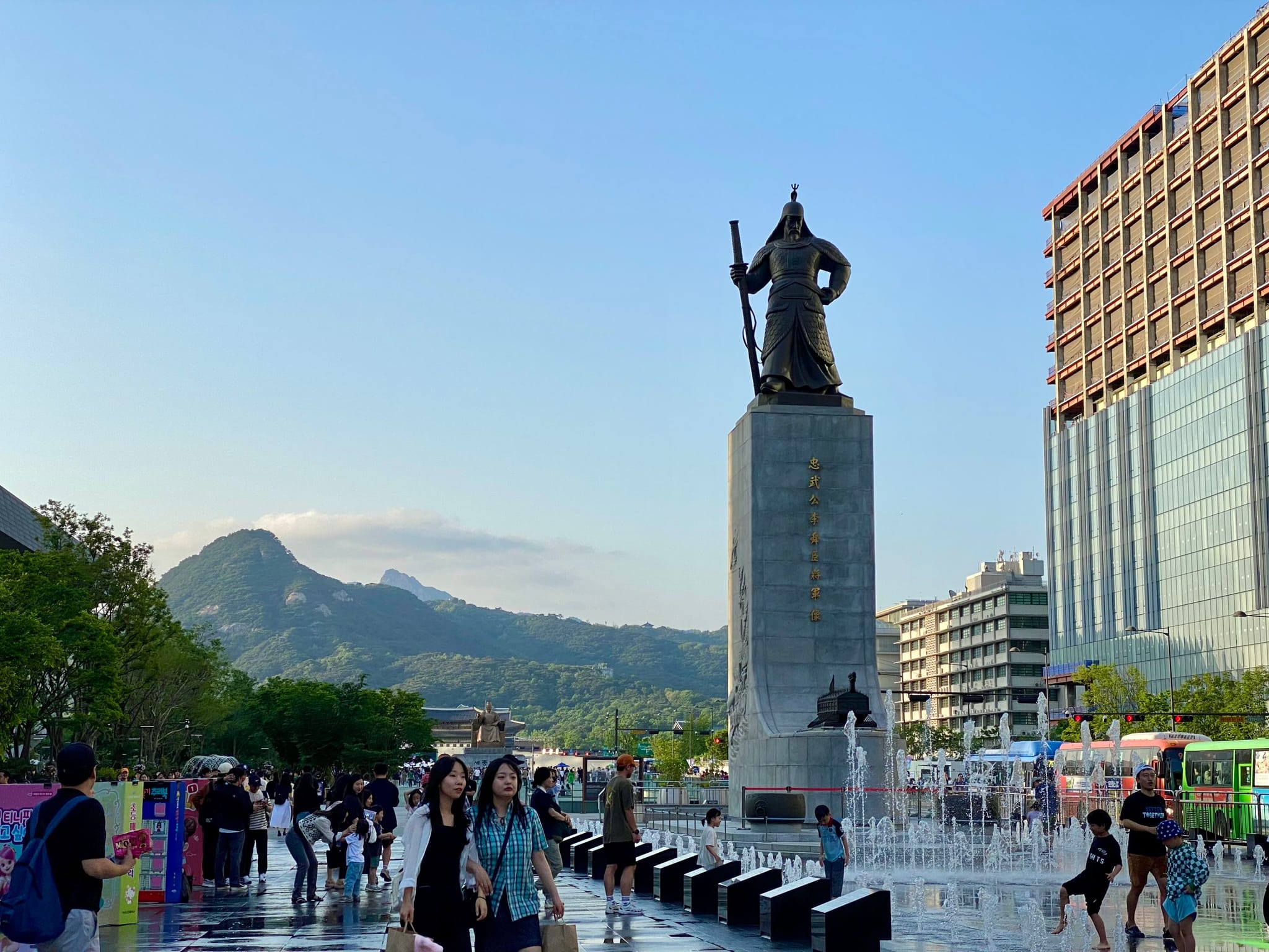 A large statue of a historical figure stands prominently in an urban plaza, surrounded by people and modern buildings, with mountains visible in the background