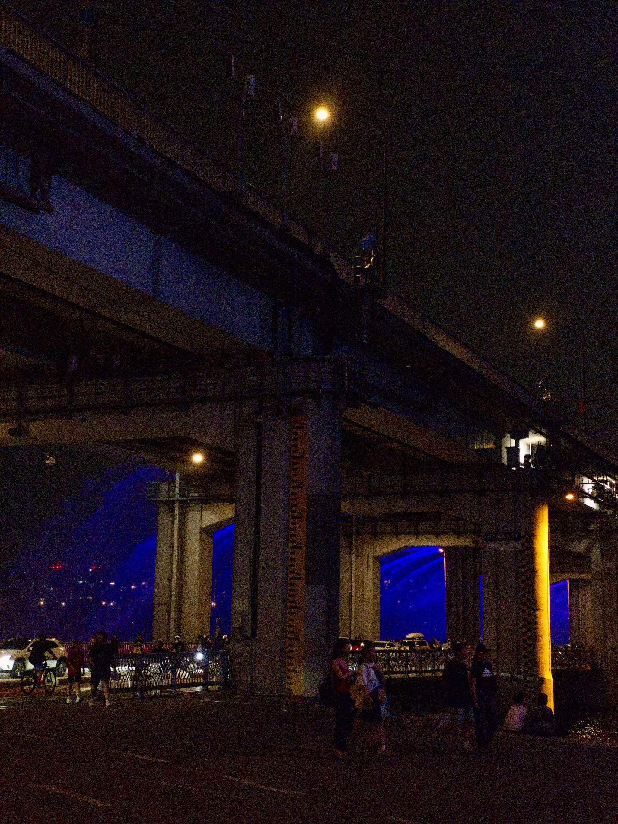 A nighttime scene featuring an elevated highway with blue and orange lighting, and people walking underneath