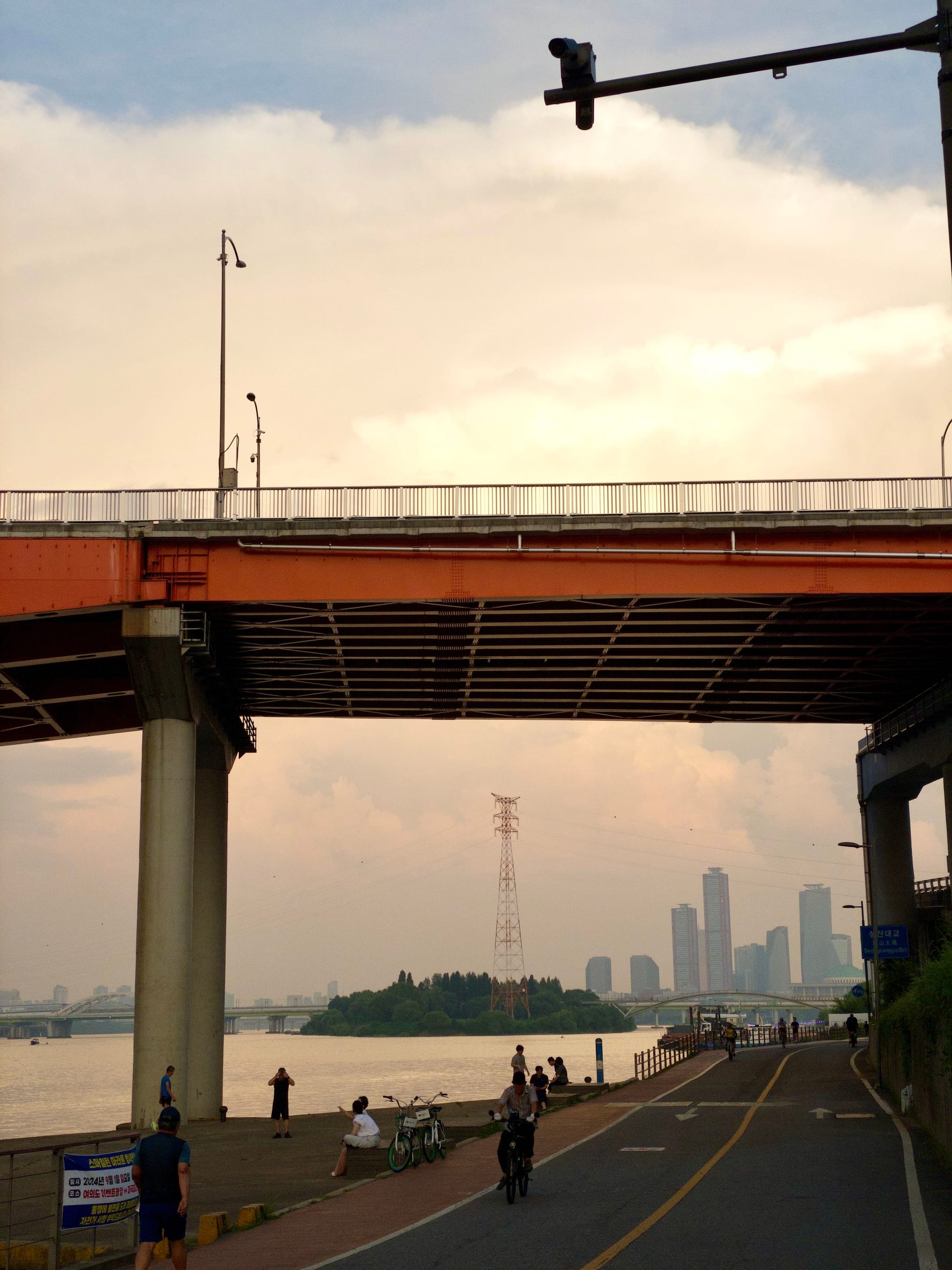 A riverside scene with a bridge overhead, people walking and cycling on a path, and a city skyline in the background under a cloudy sky