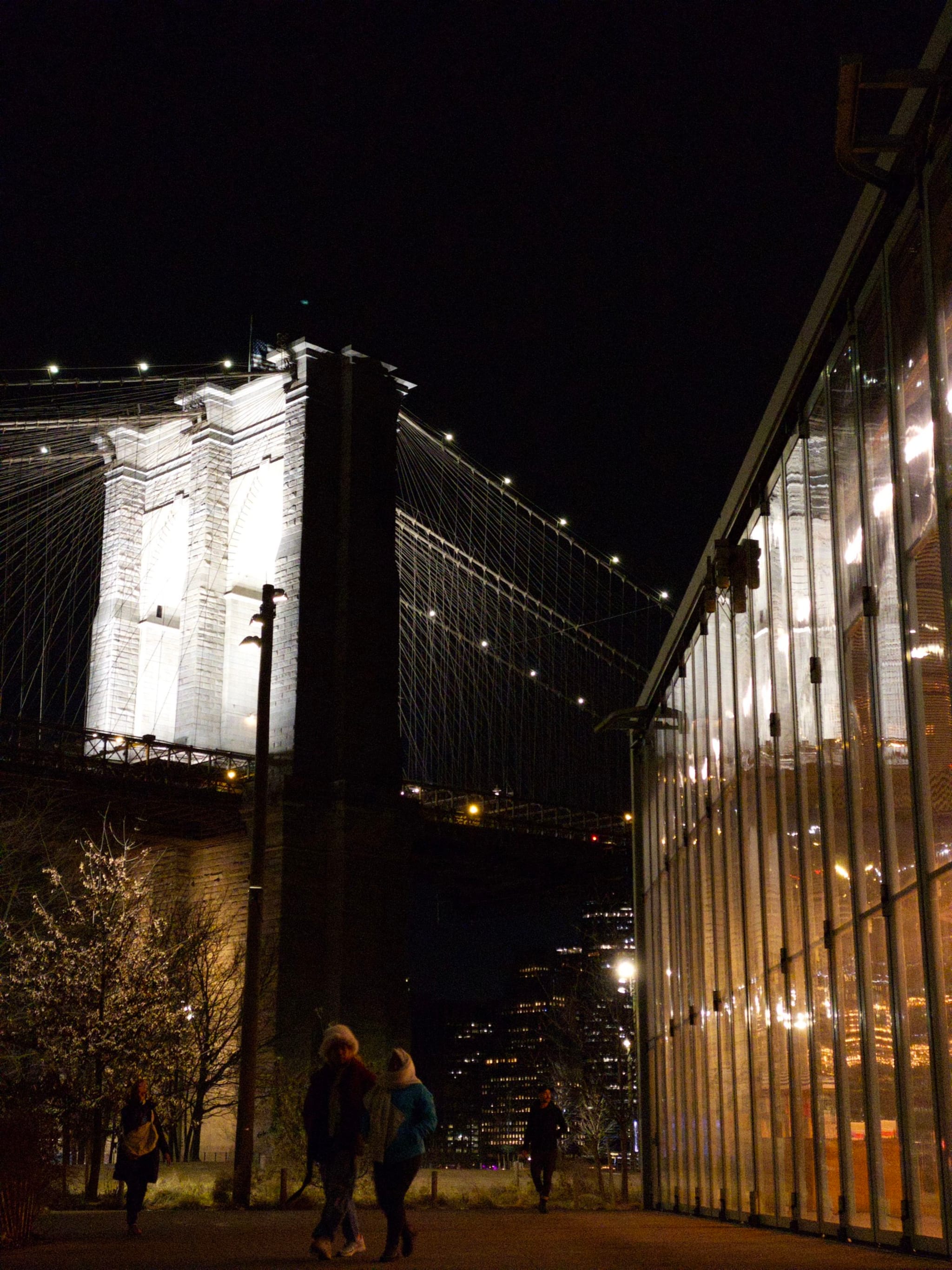 A nighttime scene featuring a brightly lit bridge with suspension cables, a glass-walled building on the right, and a few people walking in the foreground