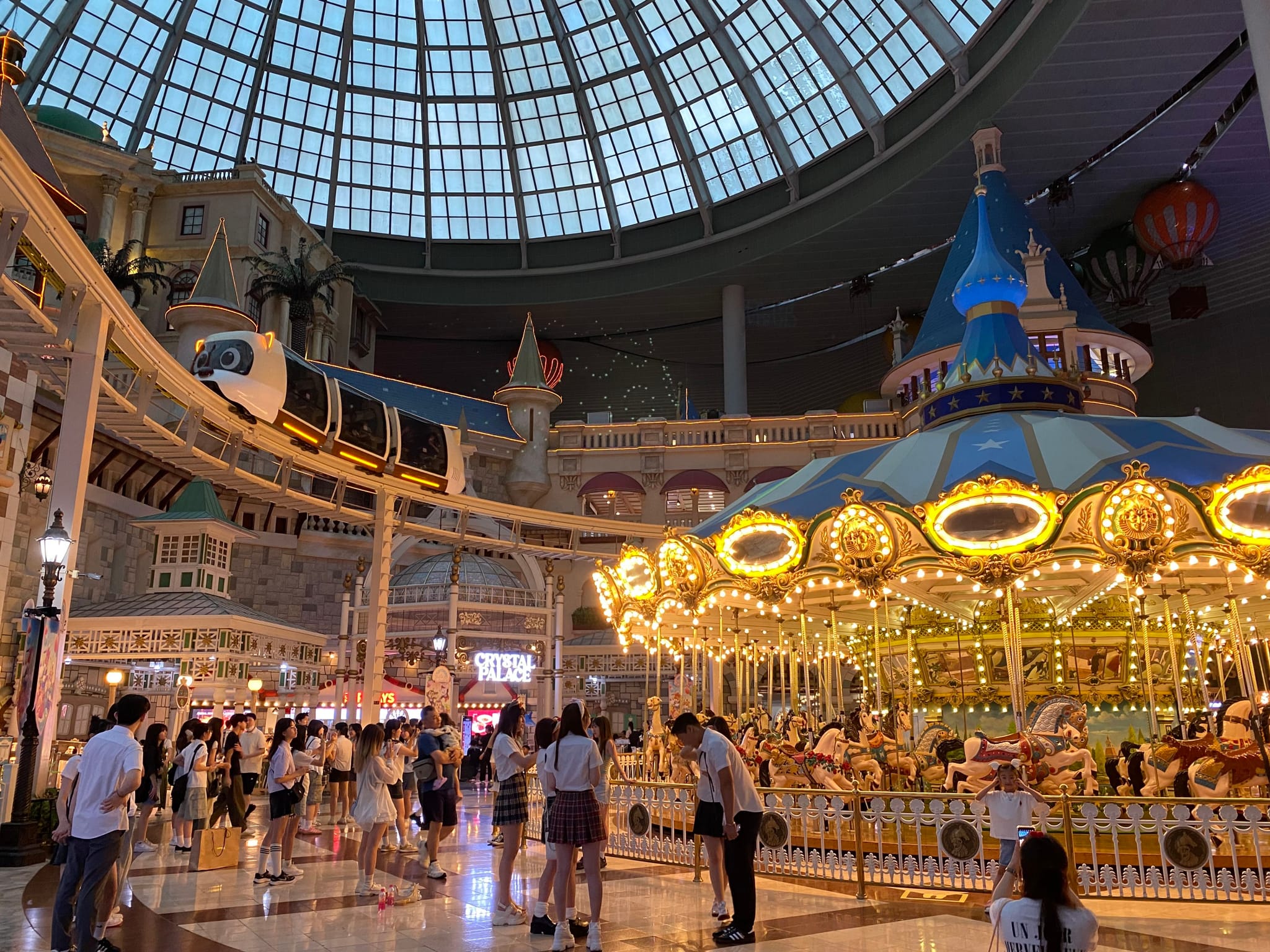 An indoor amusement park with a large carousel, a monorail track, and people walking around under a glass dome ceiling